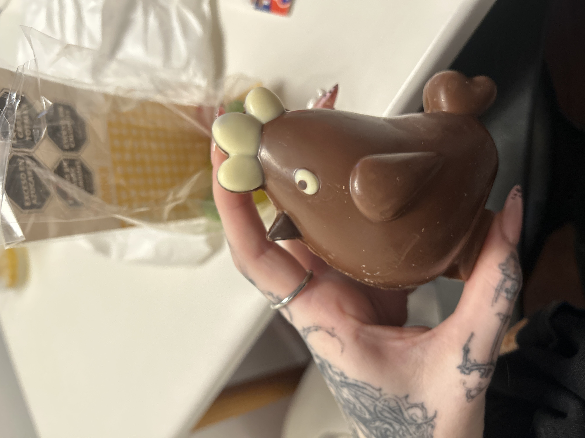 A person's hand, adorned with intricate tattoos and a silver ring, holds a novelty chocolate figure. The chocolate is shaped like a stylized bird or chick, primarily dark brown, with white chocolate details forming its eyes and a comb-like crest on its head. A small heart shape is visible on its tail.
In the background, out of focus, lies a transparent plastic bag containing food items. Several hexagonal black warning labels are visible through the plastic. These labels bear Spanish text, clearly readable on some as "EXCESO EN AZÚCARES" (Excess in Sugars) and "EXCESO EN GRASAS SATURADAS" (Excess in Saturated Fats). Through the bag, a light yellow, textured food item, possibly crackers or a waffle, can be seen. The setting appears to be indoors, likely on a light-colored table or counter. The lighting is soft and even, suggesting indoor artificial light or a bright, possibly overcast, day. The presence of these specific food warning labels is consistent with regulations in countries like Argentina.