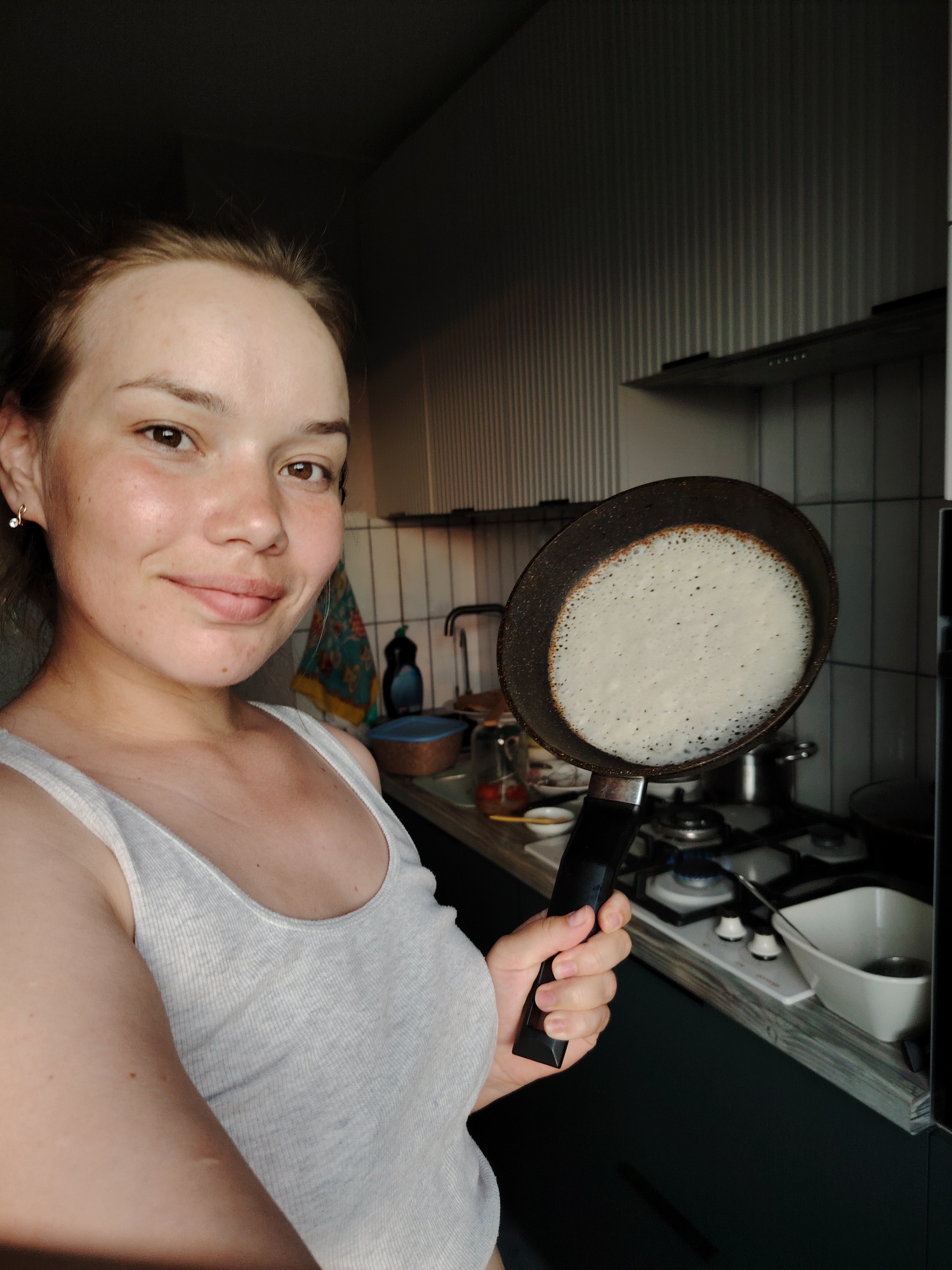 A person with fair skin and light brown hair is holding a black frying pan in their right hand in a kitchen. The pan contains a white, round object with small bubbles, possibly a pancake or a similar food item. The person is wearing a light gray ribbed tank top and is smiling slightly at the camera. The kitchen setting includes a gas stovetop with a lit burner, a counter with kitchen utensils and containers, and a tiled backsplash with a hanging towel. The lighting appears to be a mix of natural and artificial, with shadows indicating a light source from the left. The location is not discernible from the image, therefore it's impossible to know if this is Markovskiy, Russia.