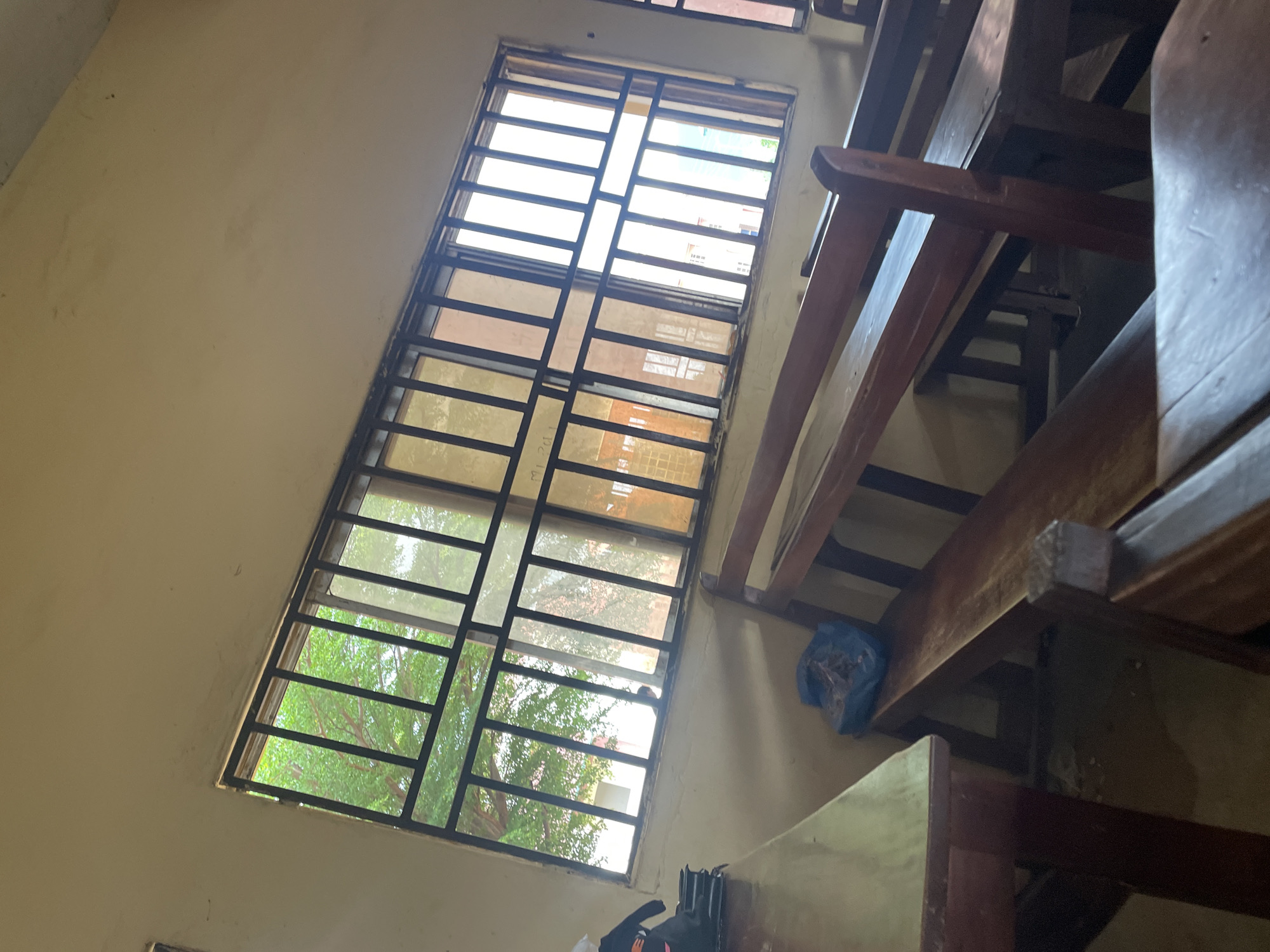 The image depicts the interior of a room, likely a classroom or assembly hall, in Lokoja, Nigeria. A prominent feature is a large window with metal bars, offering a view of lush green trees and some buildings outside. The interior is furnished with wooden benches and tables, suggesting a place for seating and study. The walls are painted a light, neutral color, and the lighting appears to be natural, coming from the window. There are no people clearly visible in the foreground, though a small figure can be seen through the window in the distance. A blue plastic bag is placed on one of the benches, and a black bag is on a table to the left. The overall impression is one of a functional, utilitarian space.