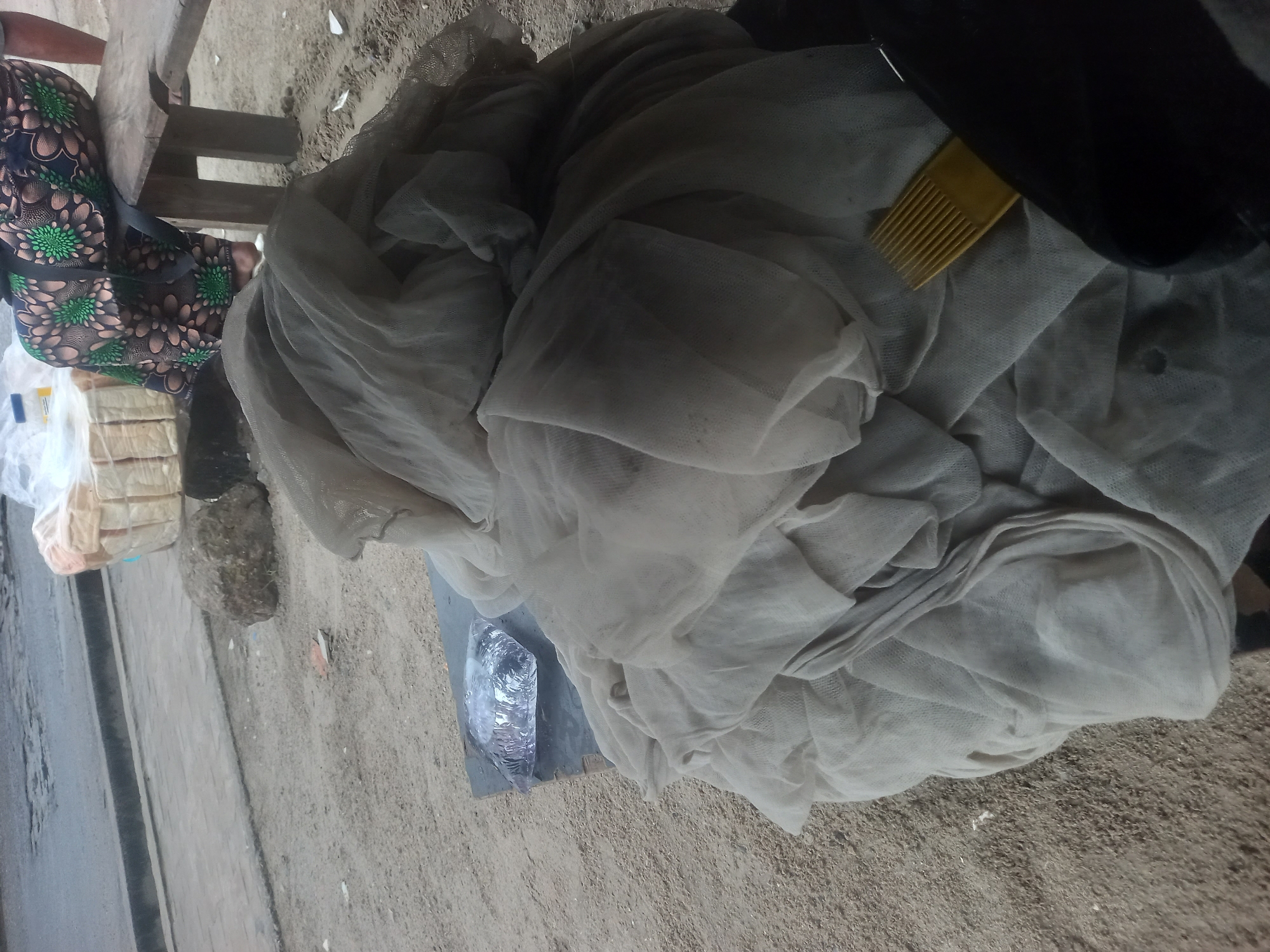 This image captures a street scene in Ilado, Nigeria, focusing on a vendor's stall. In the foreground, a large pile of light-colored netting is the primary subject, draped over a flat surface. Partially visible beneath the netting on the left is a clear plastic bag containing what appears to be fish. To the right of the netting, a yellow comb is visible, nestled within the fabric. In the background, a person wearing a vibrantly patterned skirt is seated on a wooden bench. Behind the netting and to the left, a basket of bread loaves, some wrapped in plastic, is partially obscured. A large, rough rock lies on the sandy ground between the bread and the netting. The ground itself is sandy, with some debris scattered around, and a paved pathway and asphalt road are visible in the upper portion of the image.