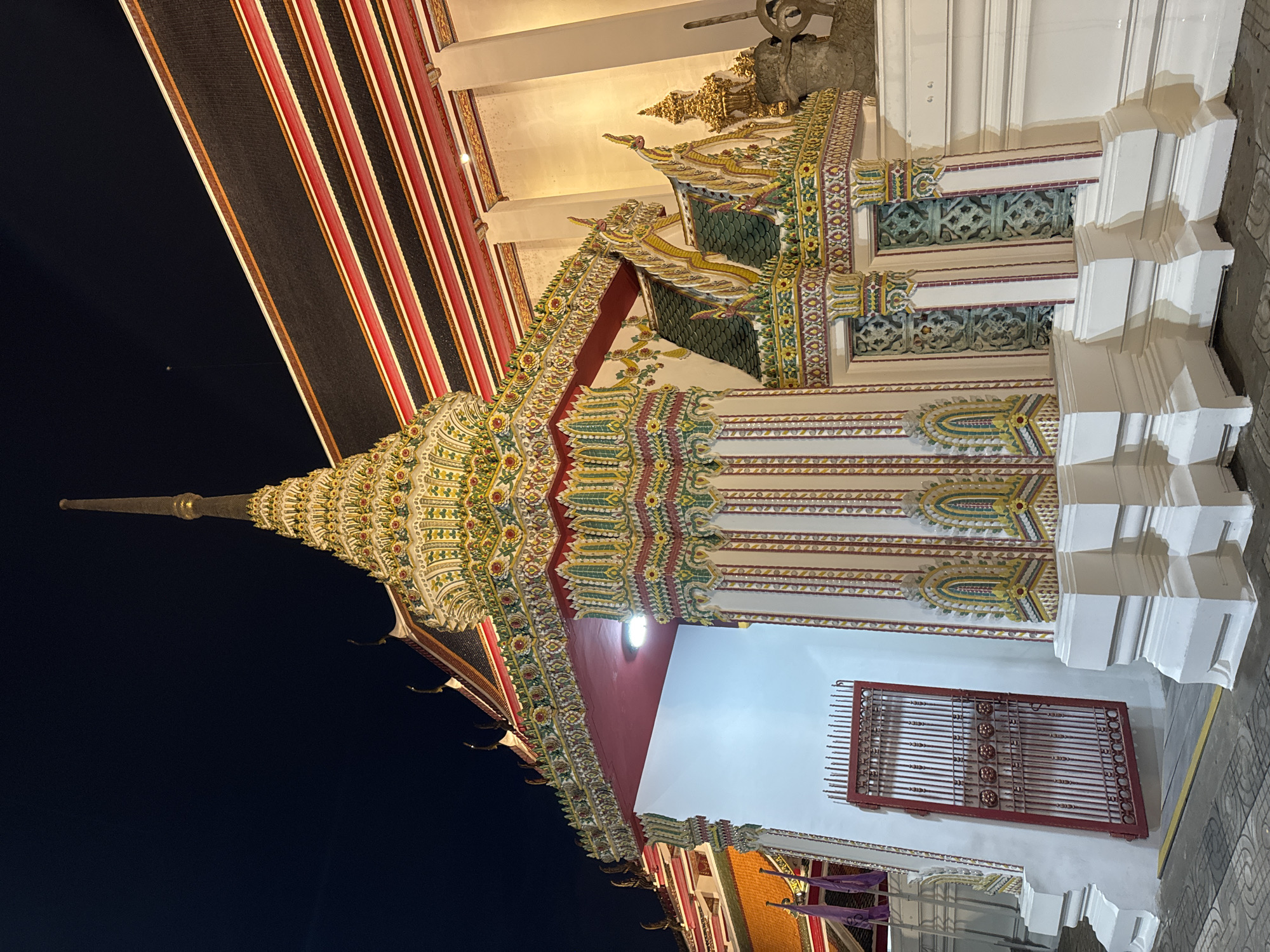 The image shows a close-up, eye-level view of the ornate exterior of a Thai temple at night. The architecture is highly decorative, featuring intricate colorful tilework, gilded spires, and multi-tiered roofs. The dominant colors are white, gold, green, red, and yellow. A tall, slender spire topped with a finial pierces the dark night sky. The building's facade is illuminated by artificial light, casting a warm glow and creating shadows. The foreground shows a patterned tiled walkway. In the background, the dark sky is devoid of stars. The image appears to be taken in Bangkok, Thailand, given the architectural style commonly found in Thai temples. No people are visible in the frame.