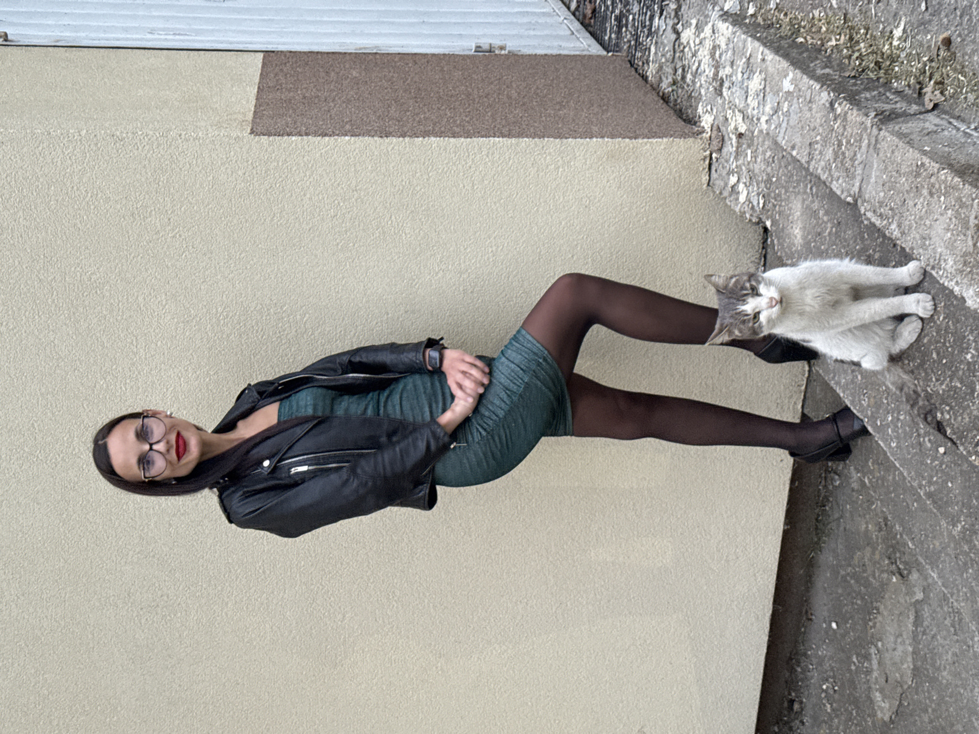 A woman in a green dress, black tights, and a black jacket is posing beside a white and gray cat sitting on a concrete step. The woman is wearing glasses and red lipstick. She is positioned in front of a beige wall with a brown stripe at the top. The setting is potentially an urban environment with visible steps and a wall that could be part of a building in Pleven, Bulgaria.