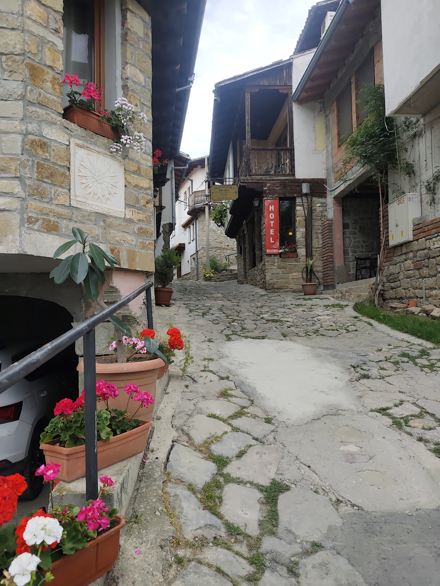 The image depicts a narrow, inclined street paved with cobblestones in the town of Unknown, Unknown. The street is lined with buildings constructed from stone and brick, some of which feature flower boxes with red and white geraniums. A hotel sign is visible on one of the buildings. A car is partially visible under an archway on the left side of the image. The overall atmosphere is characteristic of an old European village.
