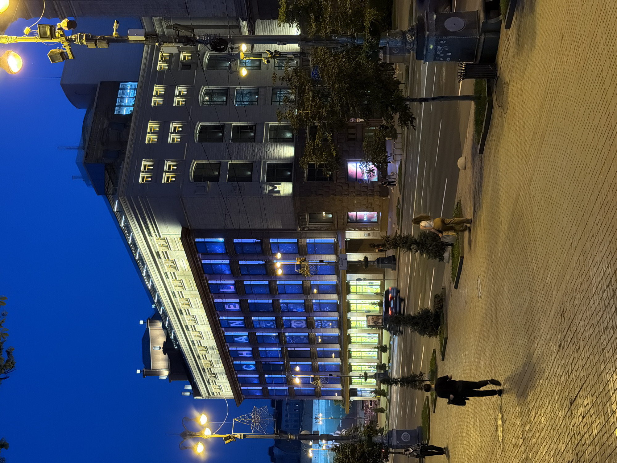 The image depicts a street scene in Kyiv, Ukraine, at dusk or early night. The primary focus is a large, multi-story building prominently displaying the word "CHANEL" in illuminated blue letters across its upper floors. The building's facade features numerous windows, some of which are lit with yellow light, while others are bathed in blue light from the display.
In the foreground, a wide, brick-paved sidewalk runs parallel to a street with multiple lanes of traffic. A few people are visible on the sidewalk. One man in a black shirt and pants walks away from the camera, carrying a bag. Further down the sidewalk, a woman in a white top and yellow pants walks towards the right side of the frame.
The street itself has a few cars, one of which is blurred by motion, indicating it is driving. Several trees are planted in rectangular beds along the sidewalk, and there are streetlights with multiple lamps illuminating the area. Decorative street lamps with starburst-like elements are also present. In the background, other buildings and urban elements are visible against a dark blue sky. The architecture suggests a commercial or upscale urban district.