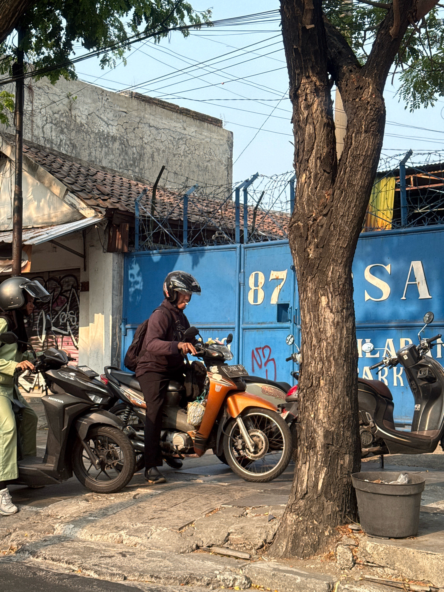 The image captures a street scene in Surabaya, Indonesia, under a clear sky with the sun casting warm light. In the foreground, a large, textured tree trunk dominates the right side of the frame, with its branches extending upwards and partially obscuring a tangle of power lines. On the left, two individuals on scooters are visible. One, wearing a green outfit and a helmet, is seated on a black Honda scooter. Next to them, another person, also helmeted, is on an orange and silver scooter, holding the handlebars and appearing to be stationary. Behind the scooters, a vibrant blue gate with large white numerals "87" and letters "SA" is prominent, suggesting it might be an entrance to a facility. Barbed wire is visible along the top of the gate. The surrounding environment includes cracked concrete pavement, a weathered wall with graffiti, and other parked scooters. A dark grey trash can is positioned at the base of the tree. The overall impression is that of a typical urban setting in Surabaya, possibly during a period of low traffic.