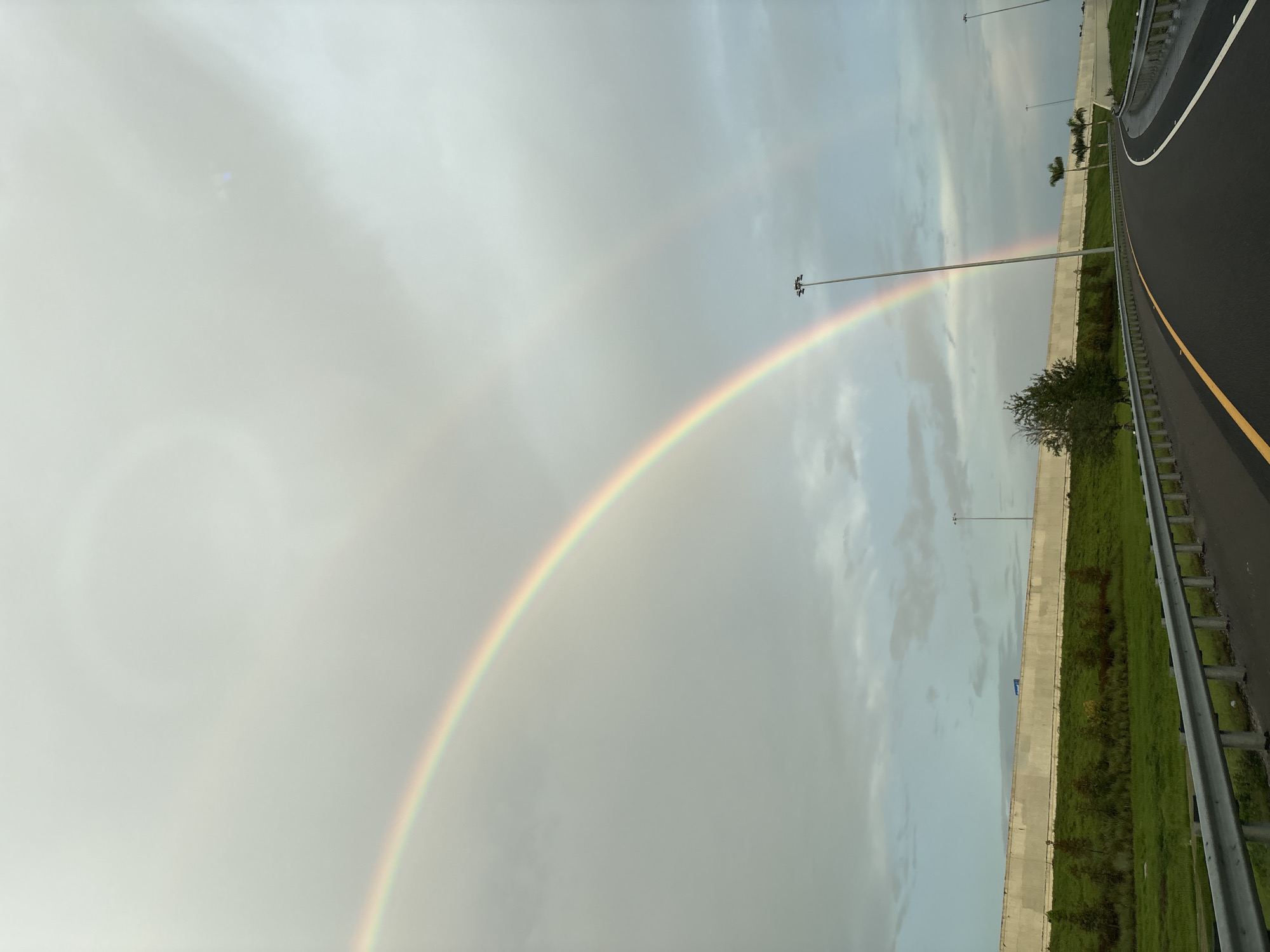 This is a photograph of a double rainbow appearing in the sky over a highway in Bradenton, Florida, United States. The sky is overcast with gray clouds, but the two distinct arcs of the rainbow are visible. The primary rainbow is brighter and more defined, while the secondary rainbow is fainter. The highway curves to the right in the foreground, with a metal guardrail lining the side. Beyond the guardrail is a grassy embankment with some scattered trees and a concrete barrier wall. Several tall light poles are visible along the highway. The scene suggests a post-rain environment.