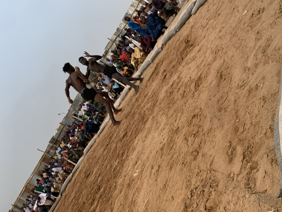 The image shows a wrestling match taking place on a sandy field in Sokoto, Nigeria. Two shirtless men wearing shorts are wrestling on the field. The man on the left is attempting to throw the man on the right to the ground. The men are surrounded by a crowd of spectators sitting along the edge of the field on sandbags. The wrestling match is taking place in an open area, most likely an outdoor arena or training field. The spectators are wearing a variety of clothing, including traditional Nigerian garb. The environment is dusty, indicating that it is likely a dry season. The sandy field is bare, with only a few footprints and the sandbags marking the perimeter of the wrestling area. The sky is clear and the weather is sunny. There are a few buildings in the background, and the atmosphere is energetic and lively.