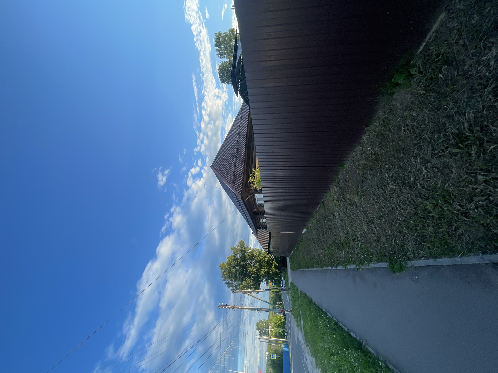 The image shows a sidewalk and a wooden house behind a dark brown metal fence in Krasnye Baki, Russia. The sky is blue with scattered clouds. There are utility poles with wires on the left side of the image, and a road with a traffic light is visible in the distance. The house appears to be a traditional wooden structure with a dark metal roof. The sidewalk is paved, and there is a grassy area between the sidewalk and the fence. The scene depicts a quiet, residential street in a rural or suburban setting.