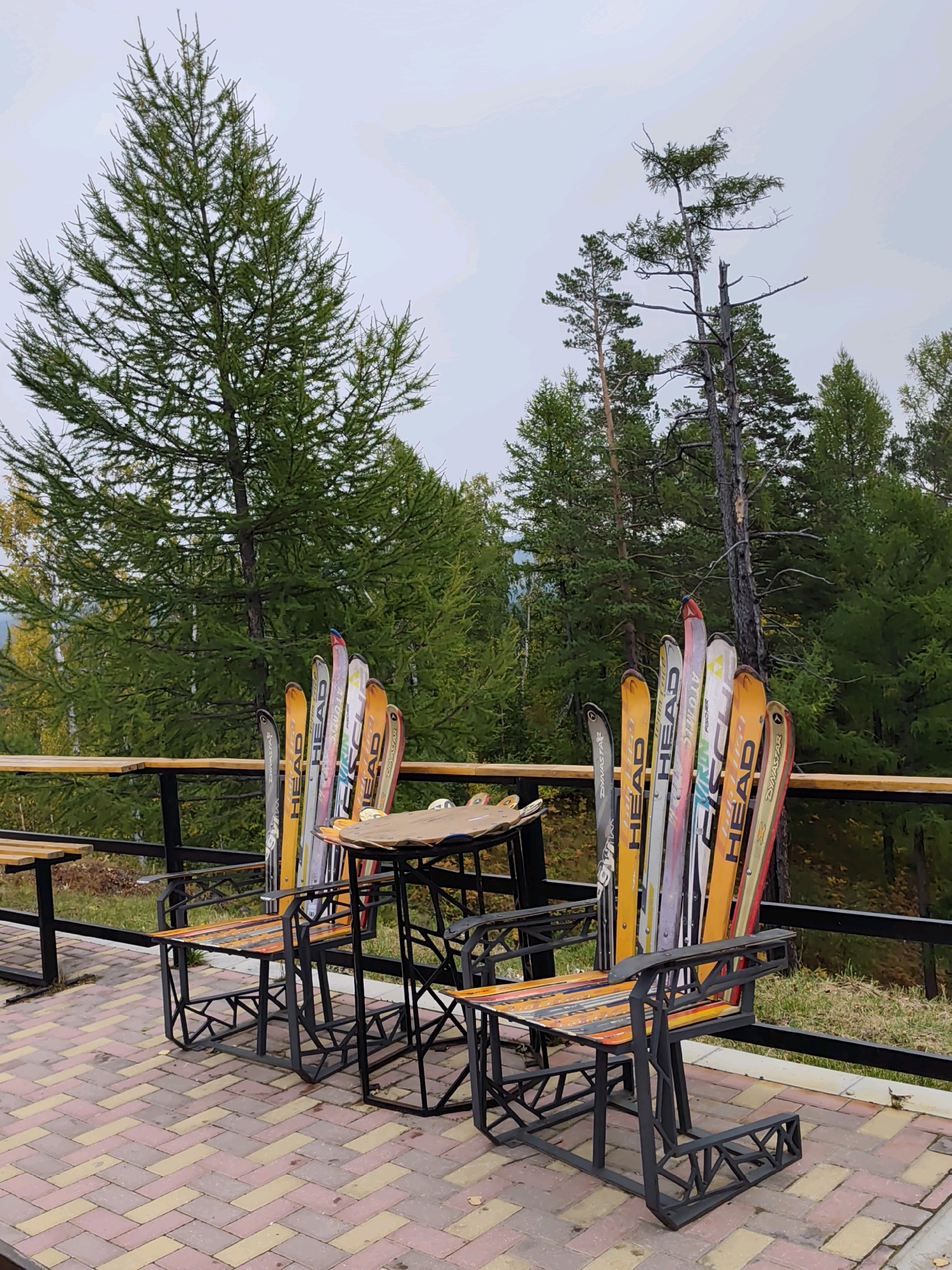 The image depicts an outdoor seating area with two chairs and a table, likely located in Nikola, Russia, given the presence of coniferous trees characteristic of Siberian forests. The chairs and table are creatively constructed from what appear to be recycled skis, arranged to form the backs of the chairs and the tabletop. The chair frames are made of black metal with intricate geometric designs. The seating surfaces of the chairs are also adorned with strips of colorful materials, possibly repurposed skis or painted wood. In the background, a dense forest of evergreen trees dominates the landscape under an overcast sky. A wooden railing runs along the edge of the seating area, separating it from a grassy slope. The ground is paved with patterned brickwork in shades of red, yellow, and beige.