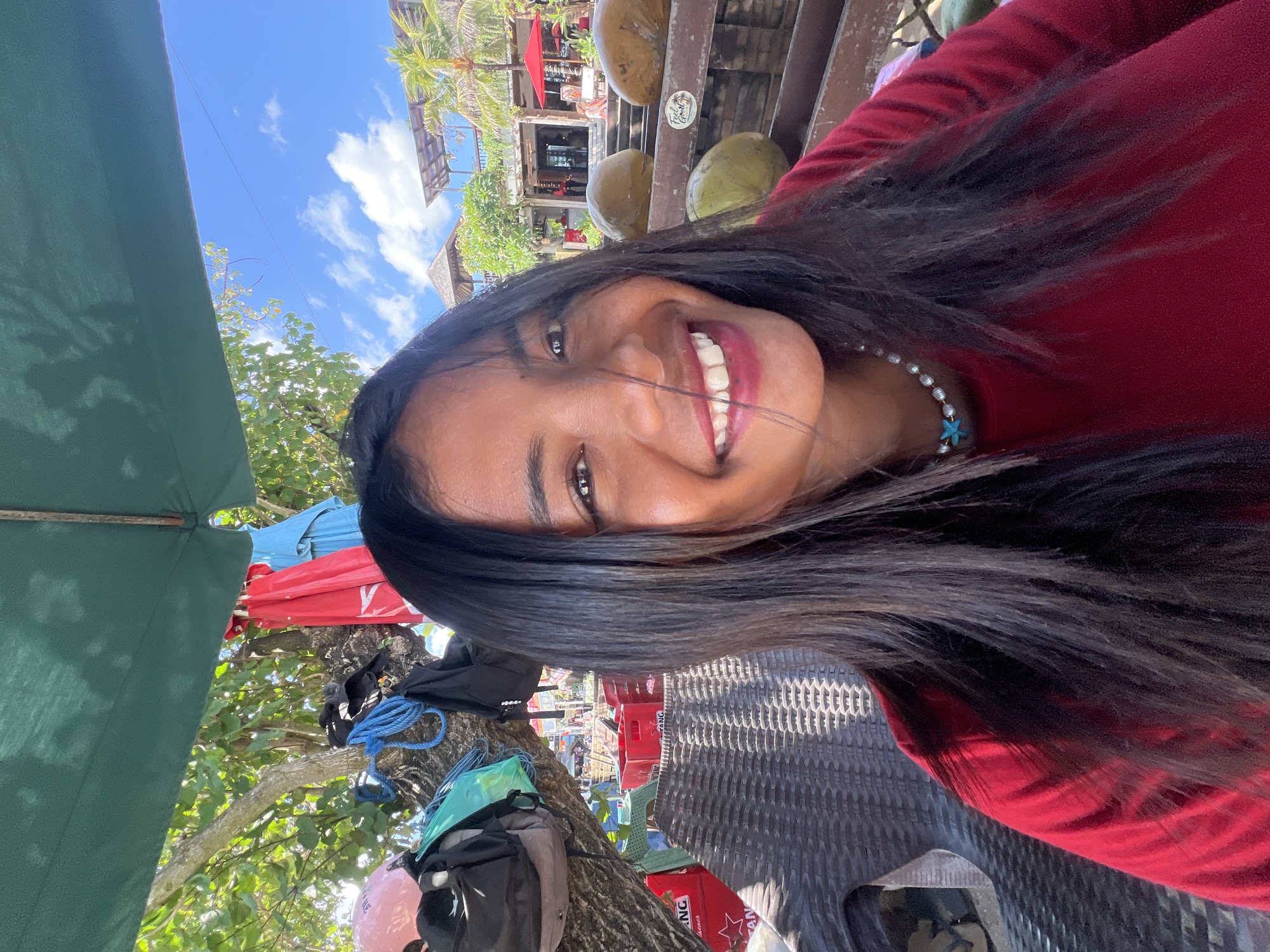In Kuta, Indonesia, a young woman with long, dark hair and a wide smile is captured in a selfie. She is wearing a red shirt and a pearl necklace with a blue star pendant. She is seated outdoors, under a green umbrella providing shade from the sun. Behind her, a tree with bags and clothing hanging from its branches is visible, along with a woven chair. Further in the background, several large coconuts rest on a wooden structure. Beyond that, buildings with wooden roofs, palm trees, and a bright blue sky with white clouds suggest a tropical environment. The scene appears to be a casual outdoor dining or resting area.