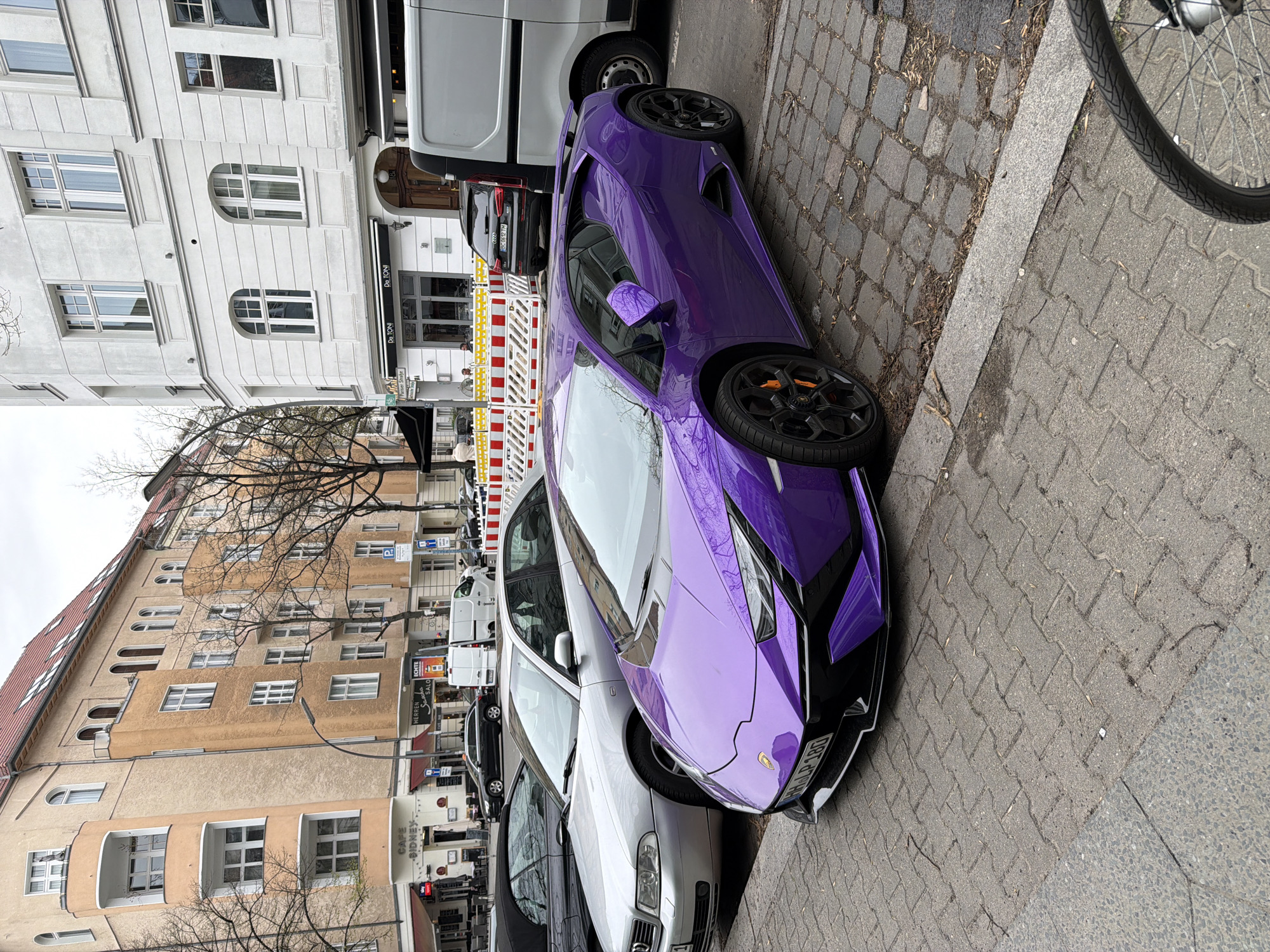A purple Lamborghini sports car is parked on a cobblestone street next to a sidewalk in Berlin, Germany. The car is parked near other vehicles, including a silver sedan and a white van. Buildings line the street, and some have storefronts. One storefront sign is visible, reading "Da TONI." A bicycle wheel is visible in the upper right corner of the image. The weather appears overcast.