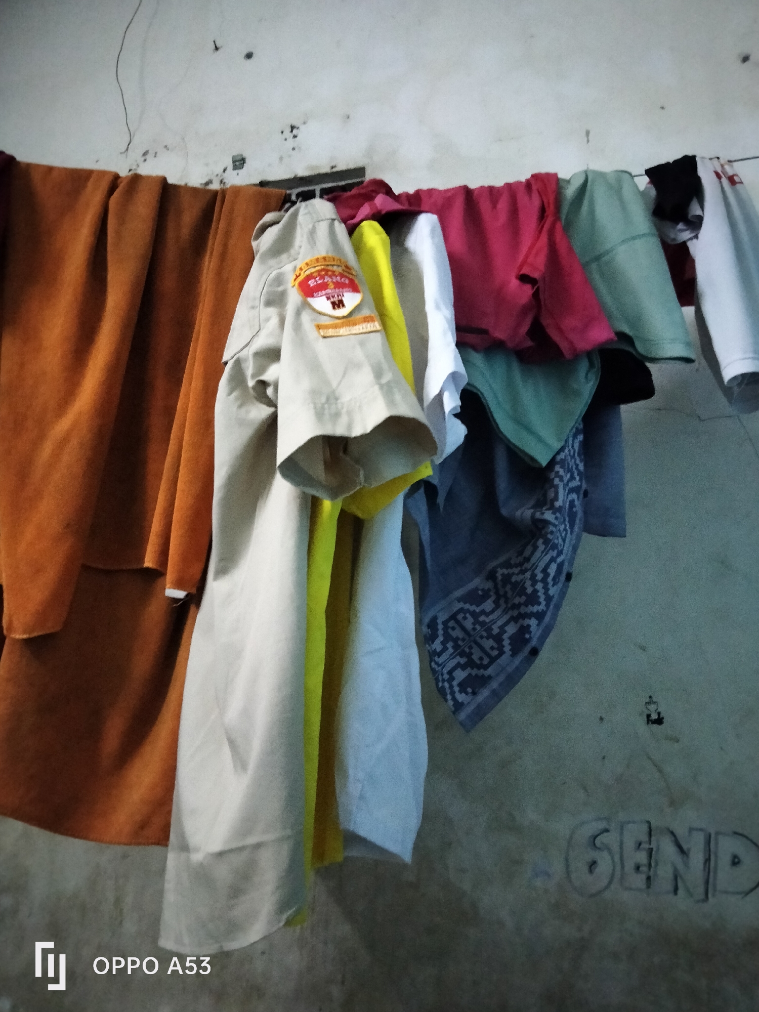 A close-up, eye-level shot captures a clothesline strung across a wall. Various articles of clothing are hanging on the line, including a tan shirt with a circular emblem on the sleeve, a bright yellow garment, a pink shirt, a green shirt, and a white shirt. A brown cloth is draped on the left side of the frame. The wall behind the clothesline appears to be concrete and shows signs of age and possible water damage. The lower right corner of the image displays the text "SEND" faintly etched into the wall, and the camera's watermark "OPPO A53" is present in the lower left corner. The scene suggests an outdoor or semi-outdoor laundry area, possibly in a residential setting. The lighting is dim, with some shadows cast by the hanging clothes. The overall impression is one of everyday life in what appears to be a warm climate. The context of Denpasar, Indonesia, would imply a tropical environment, which could influence the type of clothing and the necessity for outdoor drying.