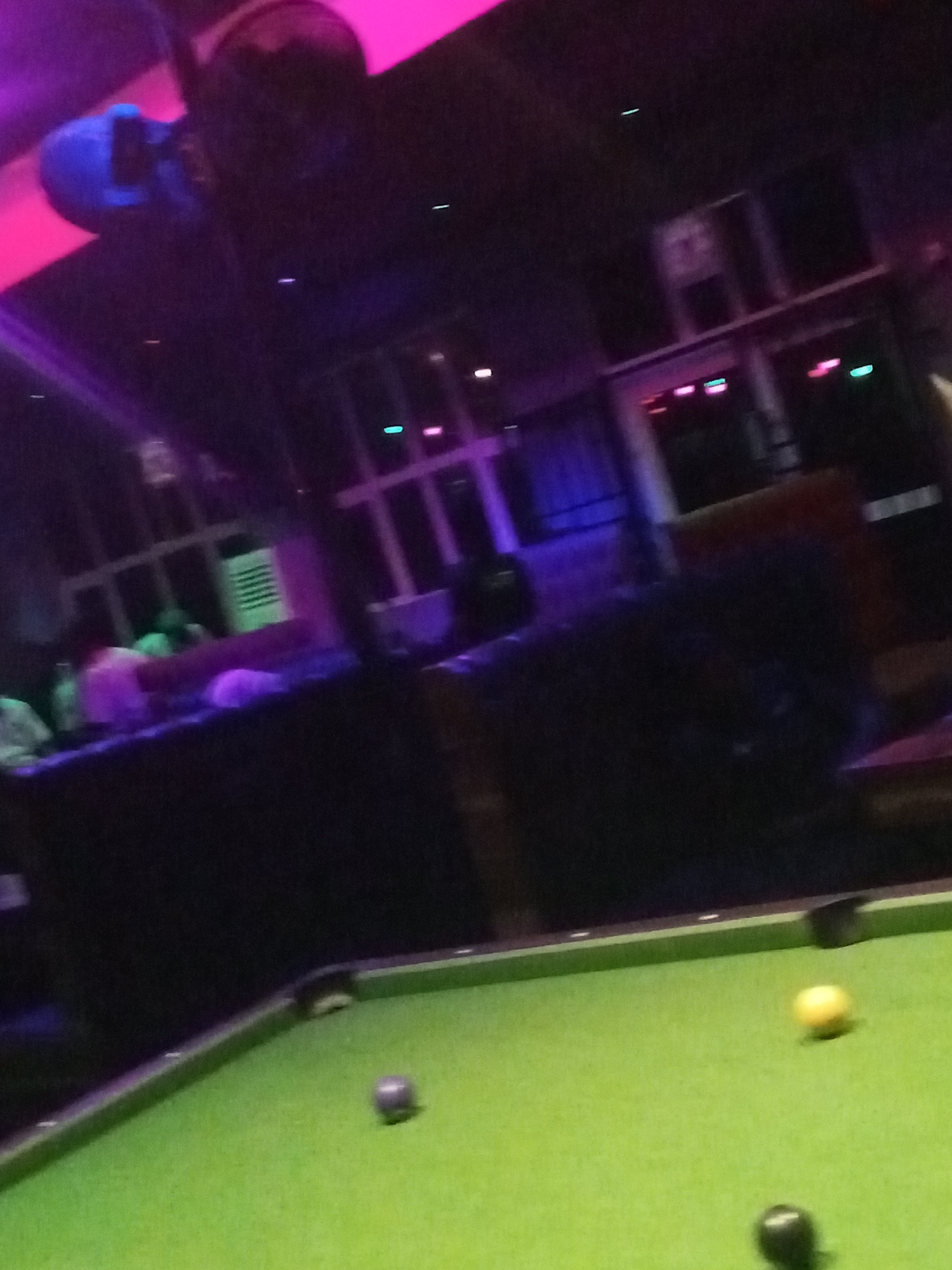 The image depicts a dimly lit interior scene, likely a bar or lounge in Lokoja, Nigeria, featuring a pool table with three visible pool balls: one yellow, one black, and one purple. The pool table is green. In the background, there are indistinct figures, likely patrons, seated at a bar under neon lights, predominantly in shades of pink, blue, and purple. The environment appears to be urban with hints of architectural elements in the back. There is a disco ball present.