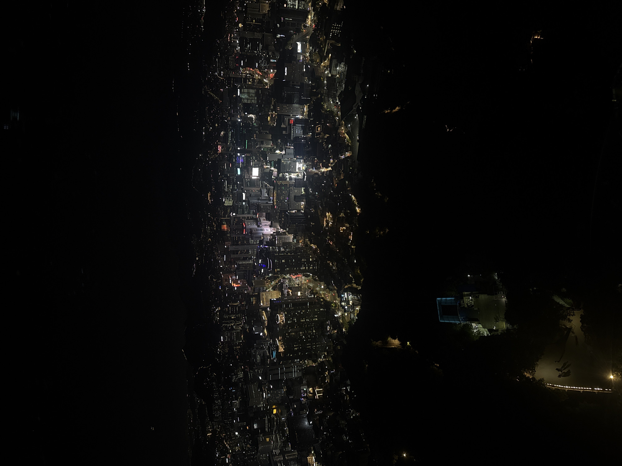 This is a nighttime aerial view of a portion of Seoul, South Korea. On the left is a densely packed urban area, identifiable by the distinct grid pattern of streets and numerous illuminated buildings. The lights suggest a mix of residential and commercial activity. To the right, a dark area likely indicates a less developed zone, potentially a park or natural landscape. There are some lights there.