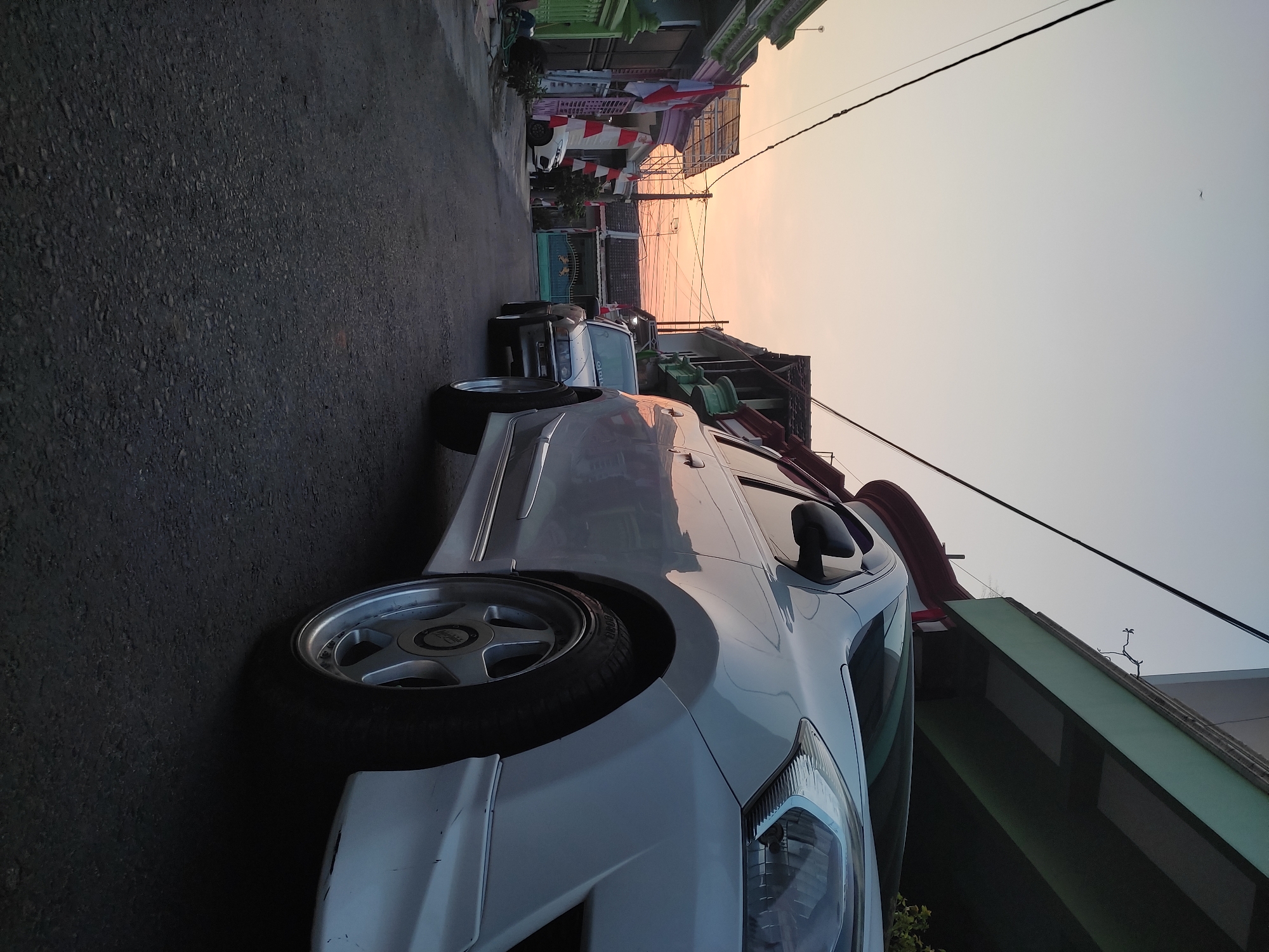 The image depicts a street scene in Ponorogo, Indonesia, during what appears to be sunset or sunrise. In the foreground, the right side of a white car is visible, angled towards the viewer. The car has a sporty appearance with custom wheels. Further down the street, another car, a silver sedan, is parked. The street is paved with asphalt and lined with houses and buildings, some adorned with Indonesian flags, suggesting a patriotic commemoration. Overhead power lines crisscross the sky, which is illuminated by soft, warm colors from the setting or rising sun. The overall atmosphere is calm and residential. There are no people visible in the image.