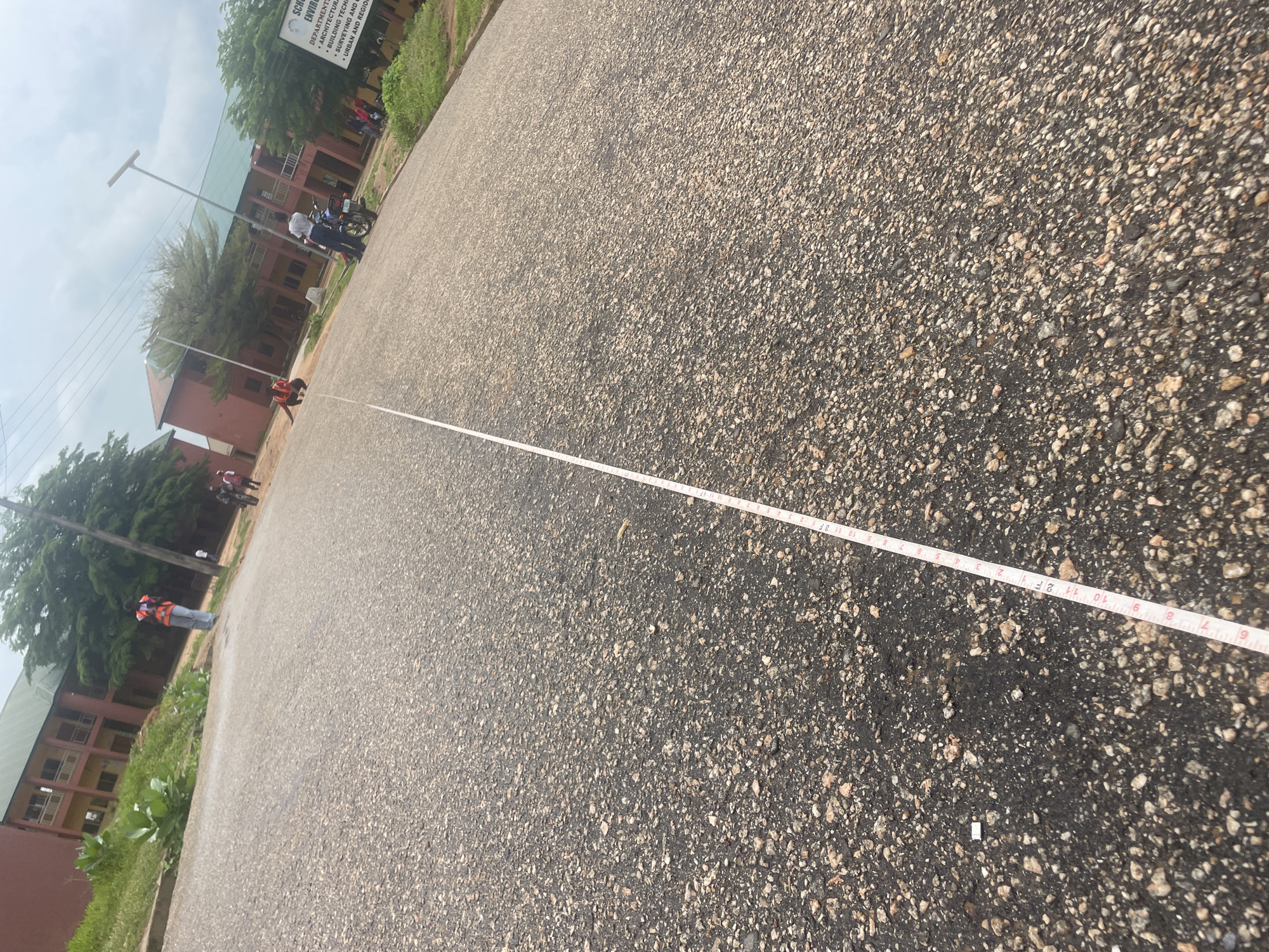 In Lokoja, Nigeria, this image captures an outdoor scene on a paved road in front of a multi-story academic building with a green roof and reddish-brown walls. The road surface is asphalt with visible aggregate. A white measuring tape, marked with black numerical increments and lines, is laid out on the road, extending from the foreground towards the middle ground.
Several individuals are present. In the foreground, a person wearing an orange safety vest and blue jeans is visible, appearing to be involved with the measuring tape. Further down the road, near the building, another individual in a white shirt stands beside a motorcycle. Two other individuals are seen on motorcycles in the middle ground, and another person in an orange vest is near the building.
The environment includes grassy areas and trees bordering the road. A signpost is visible on the right side of the image, which appears to list departments or schools, with text such as "SCHOOL OF ENVIRONM," "DEPARTMENT OF," "ARCHITECTURAL SCIENC," "BUILDING TECHNOL," "SURVEYING AND GE." and "URBAN AND REGION." The sky is overcast with clouds. The perspective is from a low angle, looking up the road.