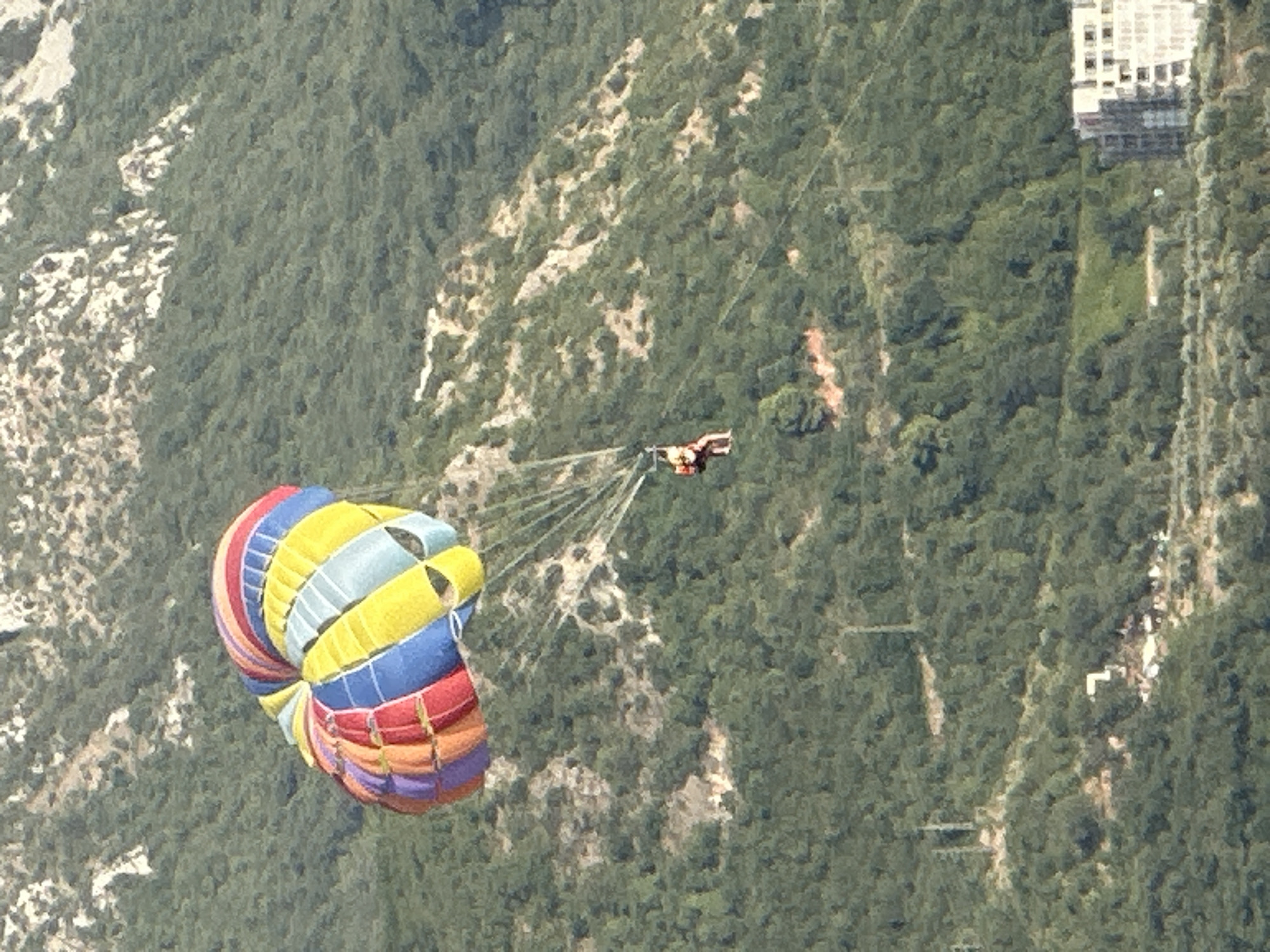 An aerial view captures a person parasailing over the verdant, mountainous terrain near Boreti, Montenegro. The parasailer, suspended beneath a vibrant, multi-colored parachute, is prominently positioned in the upper left quadrant of the image. The parachute features a bold pattern of red, orange, yellow, blue, and purple segments. The parasailer appears to be wearing a red and yellow harness. The surrounding environment is characterized by a steep, densely forested mountainside, with patches of exposed rock and sparse vegetation. In the lower right corner of the frame, a modern, multi-story building with a white facade and numerous windows is visible, suggesting proximity to a developed area. The overall scene depicts an outdoor recreational activity against a backdrop of natural beauty and distant man-made structures.