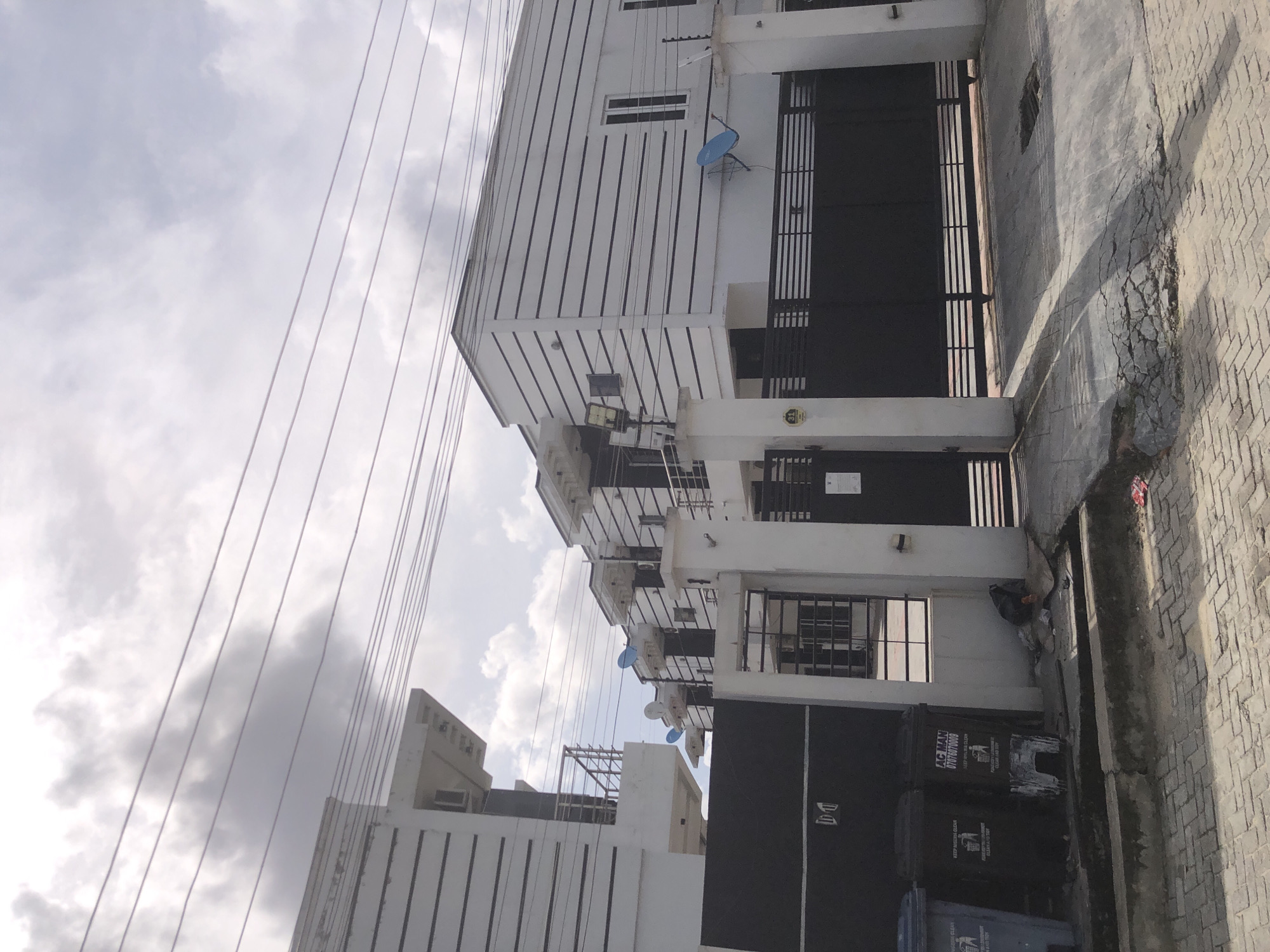 The image captures a static daytime scene in Ado, Nigeria, featuring a row of modern, multi-story residential buildings under an overcast sky. Numerous overhead power lines crisscross the upper portion of the frame, extending across the sky and in front of the buildings.
The buildings themselves are predominantly white with dark vertical accents or shutters, giving them a sleek, contemporary appearance. Each floor appears to have balconies with dark railings, and several blue satellite dishes are affixed to the facades. The ground floor of the buildings features large, dark metal gates, possibly for vehicle access or main entrances. Attached to the buildings are various outdoor light fixtures and what appear to be air conditioning units.
At street level, the foreground reveals a paved area, likely a sidewalk or a portion of the road, constructed with brick pavers. A noticeable crack runs along the edge of the pavement, suggesting wear and tear. Several dark waste bins are positioned along the wall of one of the buildings. Text is visible on two of these bins: one reads "AC MAN 07076070000," indicating a service provider and contact number. Another bin displays "KEEP WASTE CLEAN" and "CLEAN AS YOU THROW," suggesting local waste management guidelines. The weather appears cloudy or overcast, with no direct sunlight casting sharp shadows, contributing to a muted light quality. No people or explicit activity are visible in the scene.
