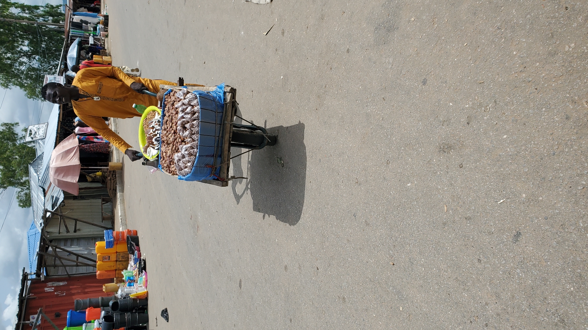 In Jalingo, Nigeria, a man pushes a wheelbarrow filled with nuts. He is wearing a yellow tunic. The wheelbarrow is covered with a blue tarp. The man is standing on a paved road with a market in the background. The market appears to be set up in an outdoor area, with several stalls and vendors visible. The sky is cloudy.