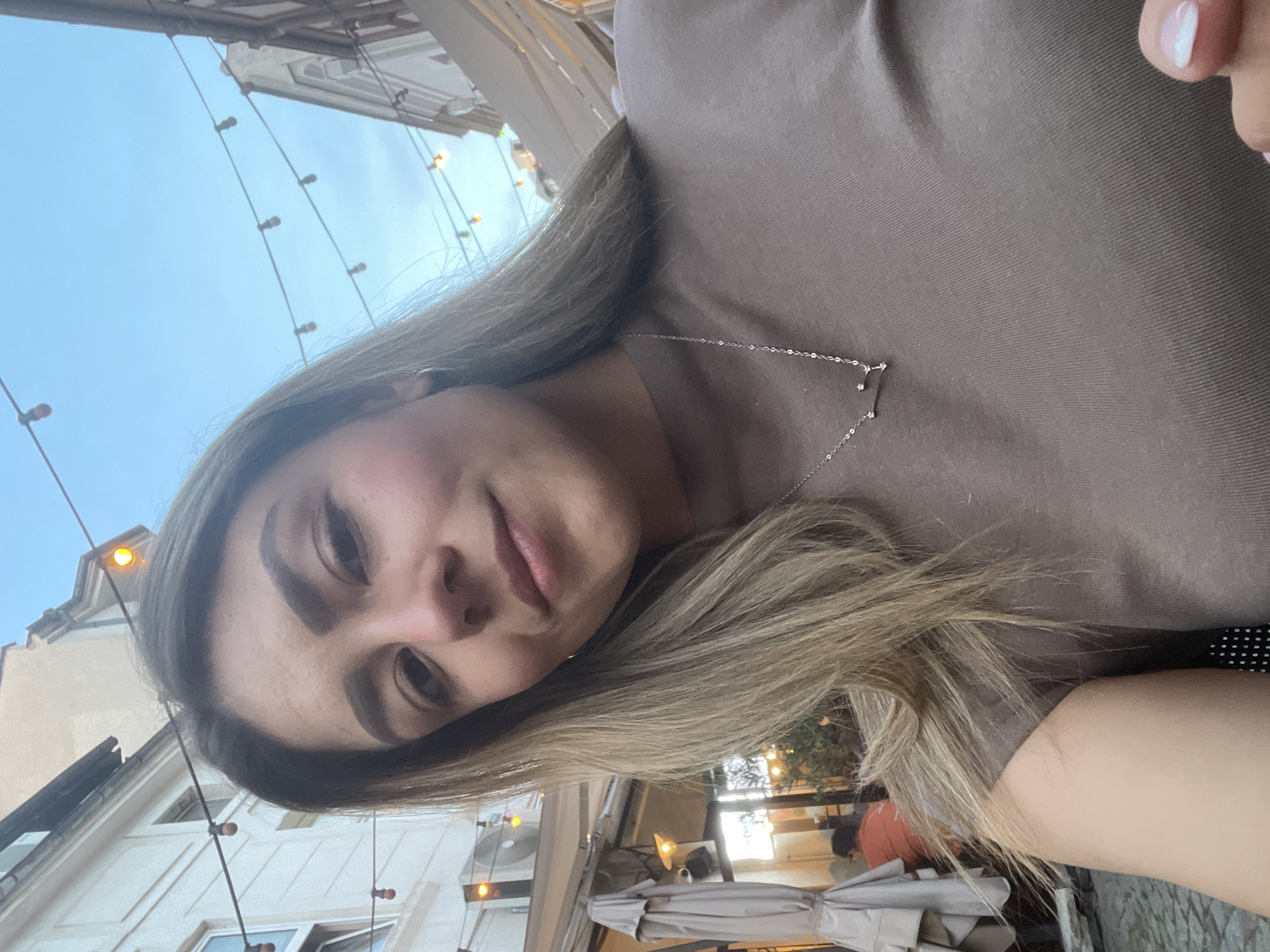 A young woman with long, blonde hair is taking a selfie outdoors. She is wearing a brown t-shirt and a silver necklace. The background shows a street scene with buildings, string lights, and outdoor seating. The lighting suggests it is either dusk or dawn. The woman is smiling slightly.