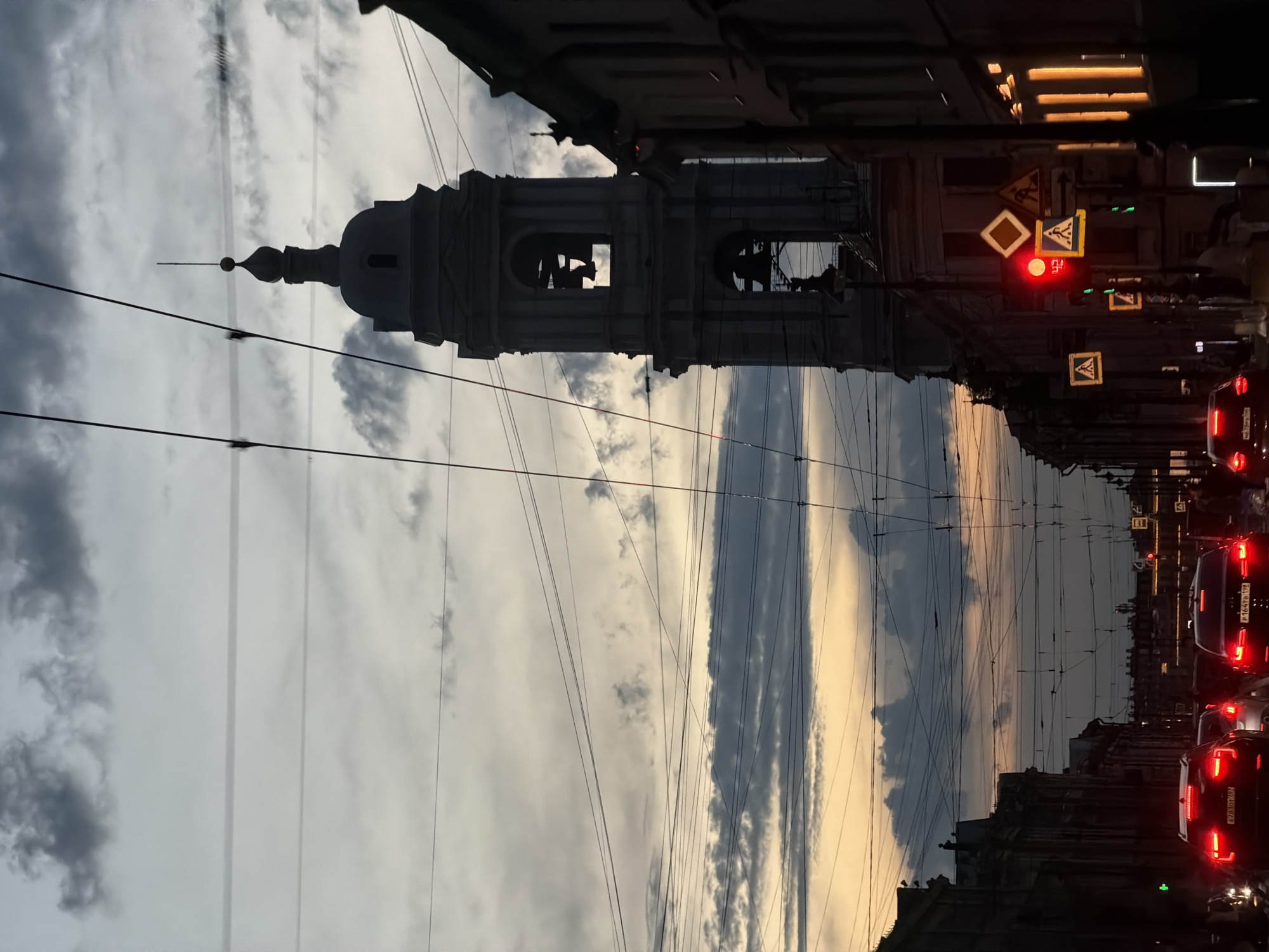 This is a photograph taken at dusk in Saint Petersburg, Russia. The image captures a view down a city street lined with buildings and overhead wires. A prominent bell tower, part of a church or cathedral, stands silhouetted against a dramatic cloudy sky that features streaks of orange and yellow from the setting sun. Several cars are visible on the street, with their taillights illuminated. Traffic signals, including a red light, are also present. The overall atmosphere is one of an urban evening scene with architectural and atmospheric elements.