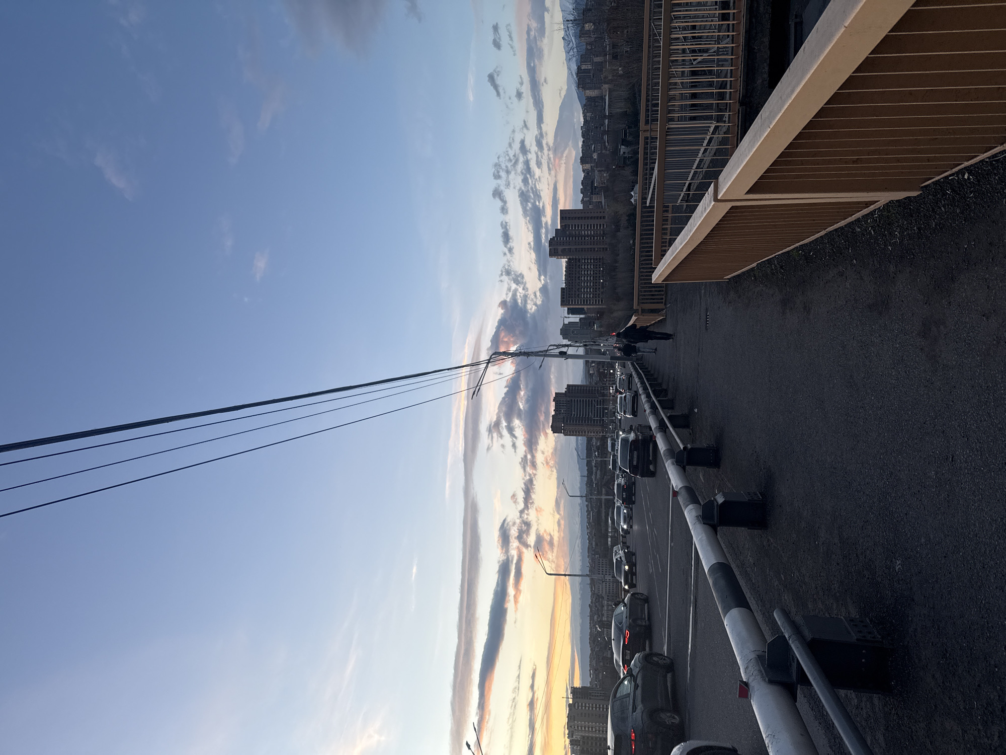 The image displays an urban landscape at what appears to be either sunrise or sunset, characterized by a dramatic sky and city structures. The perspective is from an elevated vantage point, possibly a bridge or overpass.
In the upper portion of the frame, a vast sky dominates, transitioning from a clear, light blue at the top left to warmer hues of orange, pink, and pale yellow closer to the horizon on the left and center. Scattered clouds are present, ranging from wispy white formations to more substantial, darker grey and purple clouds, particularly along the mid-horizon.
The horizon reveals a sprawling city with numerous multi-story buildings, casting a dense silhouette against the colorful sky. In the far distance, beyond the city, tall mountains or hills are visible, with some peaks appearing lighter, possibly indicating snow or distinct rock formations.
On the right side of the image, a sturdy, light-colored barrier or railing runs along the edge, appearing to be part of a pedestrian walkway or the structure of the elevated platform. Beneath this barrier, there is a dark paved surface. Further to the right, a darker, more intricate metal railing is visible. A single person, dressed in dark clothing, is walking on this paved path towards the background.
The central and left portions of the image feature a road, possibly a multi-lane highway or main street, which curves gently to the left. It is lined with numerous cars, mostly sedans and SUVs, suggesting traffic or parking. These vehicles are oriented to move away from the viewer. A distinct black and white striped barrier or divider runs along the left edge of this road, separating it from the paved area or another section. Overhead, multiple dark utility lines stretch across the sky from the left side of the frame.
The overall lighting suggests either early morning or late evening, with the warm glow on the clouds indicating the position of the sun near the horizon, contributing to a serene yet bustling atmosphere. No discernible text is visible in the image.