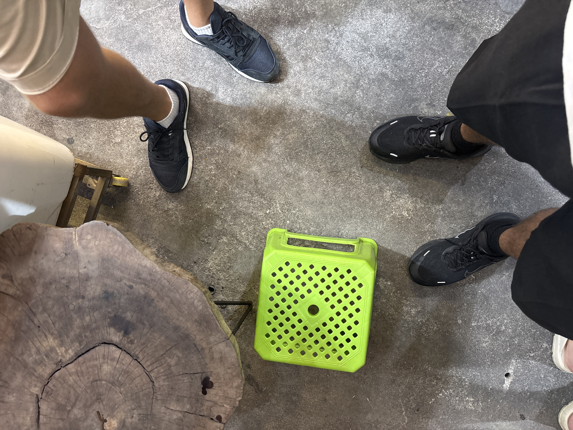 The image presents an overhead view of four individuals' lower legs and feet on a rough, textured concrete floor. In the upper left quadrant, a large, irregularly circular wooden slab, exhibiting distinct tree rings and a central crack, functions as a table surface. Below this, positioned centrally, is a lime green plastic stool with a perforated square top and an integrated handle on one side.
In the upper right, a person (Person 1) wears white shorts and navy blue athletic shoes with white soles and white ankle socks; a portion of their calf is visible. Adjacent to Person 1, in the middle right, another person (Person 2) wears similar navy blue athletic shoes with white soles and white ankle socks, with only their foot and lower ankle visible. A third individual (Person 3) occupies the lower central and right portion of the frame, wearing black shorts/trousers, black athletic shoes with white side accents, and black ankle socks; their lower legs and feet are fully visible. In the lower left, the tip of a light-colored shoe, possibly white or pink, indicates the presence of a fourth person (Person 4).
Partially visible in the upper right, behind Person 1's leg, is a white rectangular object resting on a yellow-wheeled metal frame, suggestive of a utility cart or mobile storage unit. The scene is illuminated evenly, casting soft shadows, and depicts a casual gathering around the table and stool. The location is in the city of Unknown, Unknown.