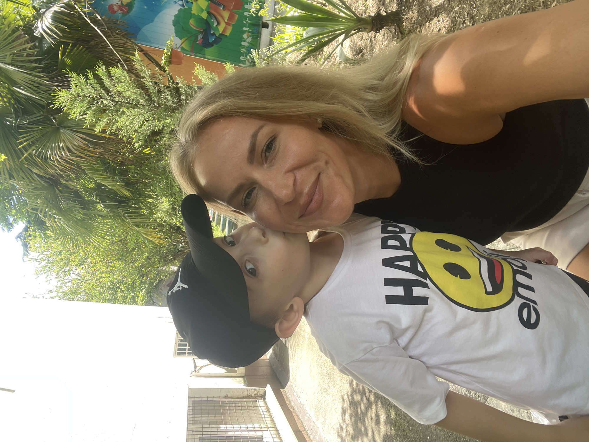 A medium shot shows a blond woman smiling directly at the camera while a young boy wearing a black baseball cap and a white t-shirt with a smiley face graphic kisses her on the cheek. The boy is on the left side of the frame, and the woman is on the right. Behind them, a white building with barred windows is visible on the left, and lush green palm trees and other foliage fill the background, suggesting a warm climate. A colorful mural depicting abstract shapes and characters is partially visible on a wall to the right. The ground is covered in dirt and gravel. The scene appears to be outdoors in a sunny, possibly tropical or subtropical location, consistent with the city of Sochi, Russia, known for its Black Sea coastline and abundant vegetation. The woman is wearing a black tank top, and the boy's t-shirt has the words "HAPPY" and "emo" printed on it. The lighting indicates it is daytime, with dappled sunlight filtering through the trees.