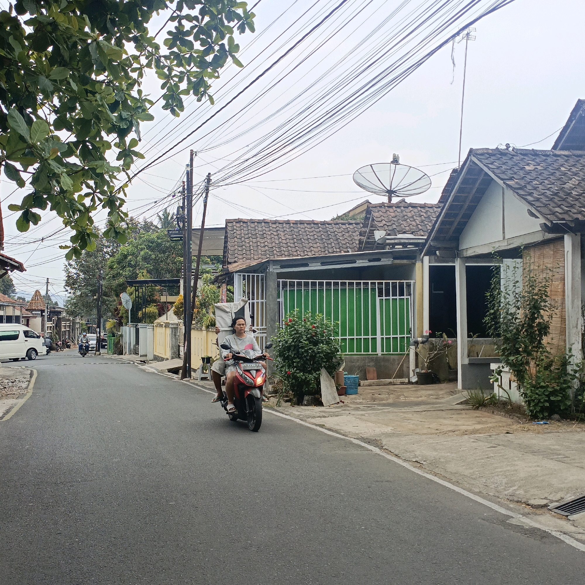 The image depicts a residential street in Salatiga, Indonesia, with a man and a woman riding a red motorcycle. The man is driving the motorcycle, and the woman is seated behind him, holding onto him. They are both wearing casual clothing. The street is paved with asphalt and has a white line marking the edge. On the right side of the street, there are houses with tiled roofs and green metal fencing. The houses appear to be single-story structures with some vegetation in front of them. The background shows more houses, trees, and a network of overhead power lines. The sky is overcast, suggesting a cloudy day. The overall impression is of a typical urban scene in a residential area of Salatiga.