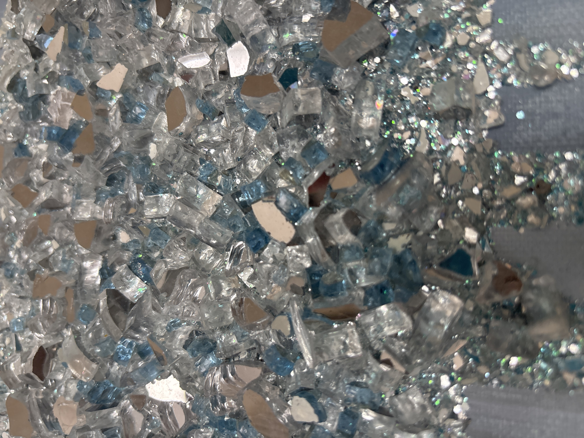 The image is a close-up, high-angle shot of a collection of broken glass fragments, some of which are tinted light blue. The fragments vary in size and shape, with sharp, irregular edges. Many pieces have reflective surfaces that catch the light, creating a shimmering effect. Interspersed with the larger glass pieces are smaller, glittering particles, possibly glitter or very fine glass chips, that add to the overall sparkle. The background is blurred and appears to be a light blue, textured surface, possibly fabric or a painted wall. The lighting is bright, emphasizing the reflective qualities of the glass. There are no people or identifiable objects other than the glass and glitter. The setting is abstract and decorative.