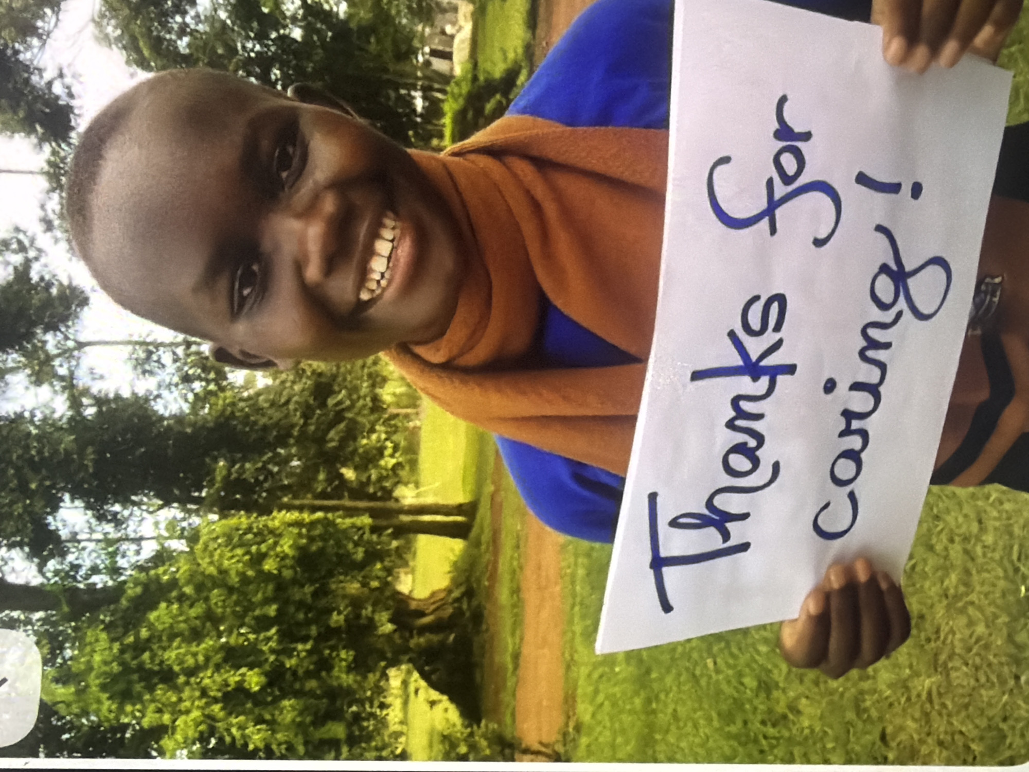 A close-up, outdoor, medium shot shows a young person with dark skin and a shaved head smiling as they hold a white sign that reads "Thanks for caring!" in blue marker. The person is wearing a blue shirt and an orange scarf. The background consists of lush green grass and trees. The scene is set in a park-like area, possibly a school or community center, in Unknown, Unknown. The lighting is natural, indicating it is daytime.