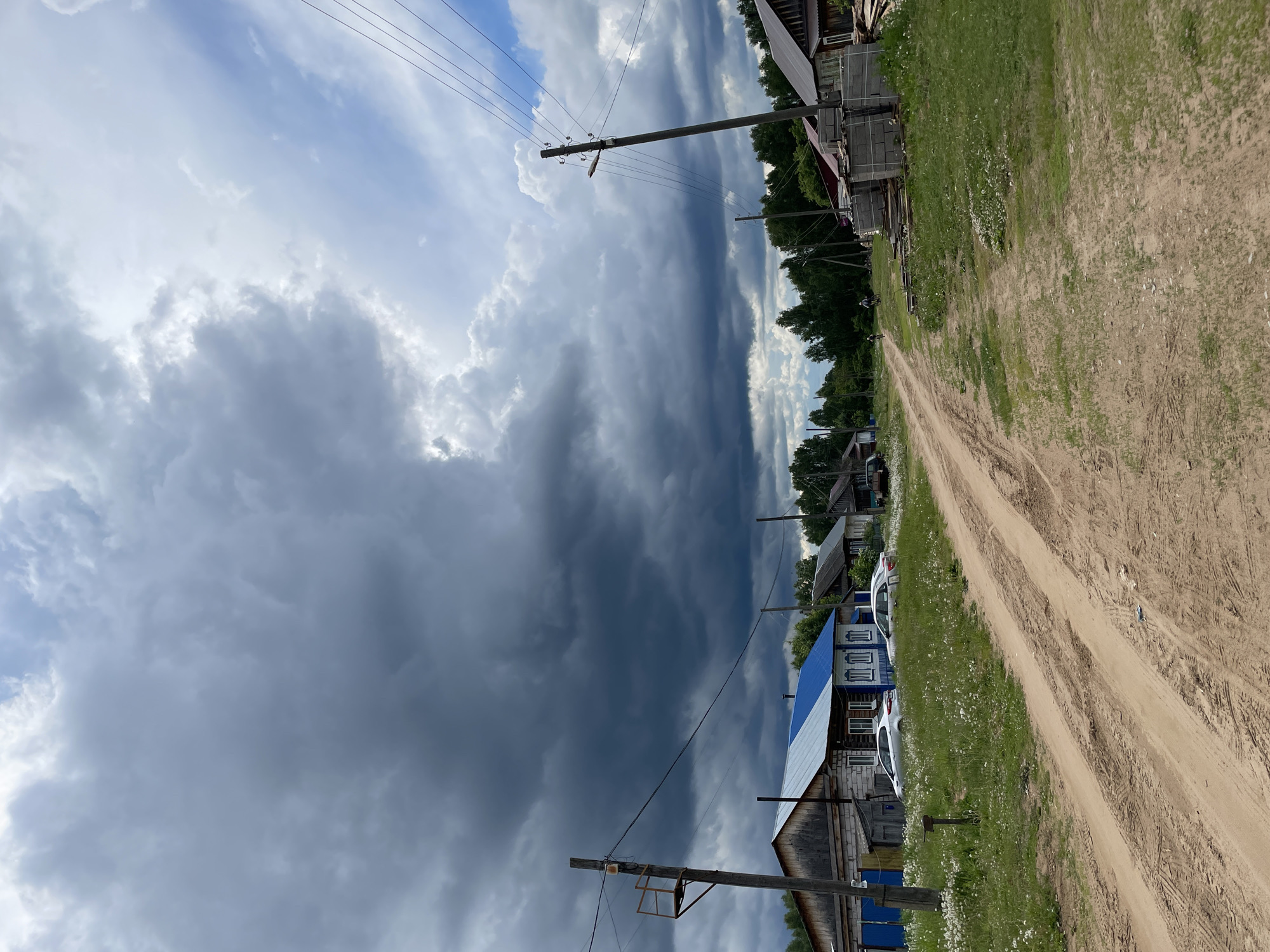 The image captures a rural dirt road in Балдейка, Russia, adjacent to simple, wooden houses. A storm is approaching, evident from the dark, ominous storm clouds filling the left half of the sky, contrasting with patches of bright sky. Power lines extend across the upper part of the image. The road's surface is dusty, showing tire tracks. Vegetation and wildflowers grow along the roadside. Several cars are parked outside the house to the left. The scene conveys a sense of impending weather in a rustic environment.