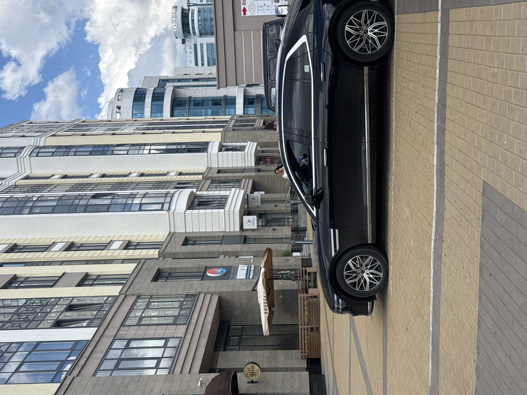 The image depicts a street scene in Yekaterinburg, Russia, with a modern building complex and a black sedan in the foreground. The building on the left has a stone facade with large windows, and signs indicating a business. A beige umbrella with the word "BROOSKY" is visible. Further down the street, a series of contemporary buildings with glass facades dominate the skyline. A few people are seen walking near the buildings. The black sedan is parked on a paved surface, with its front wheel and side profile clearly visible. The sky is partly cloudy.