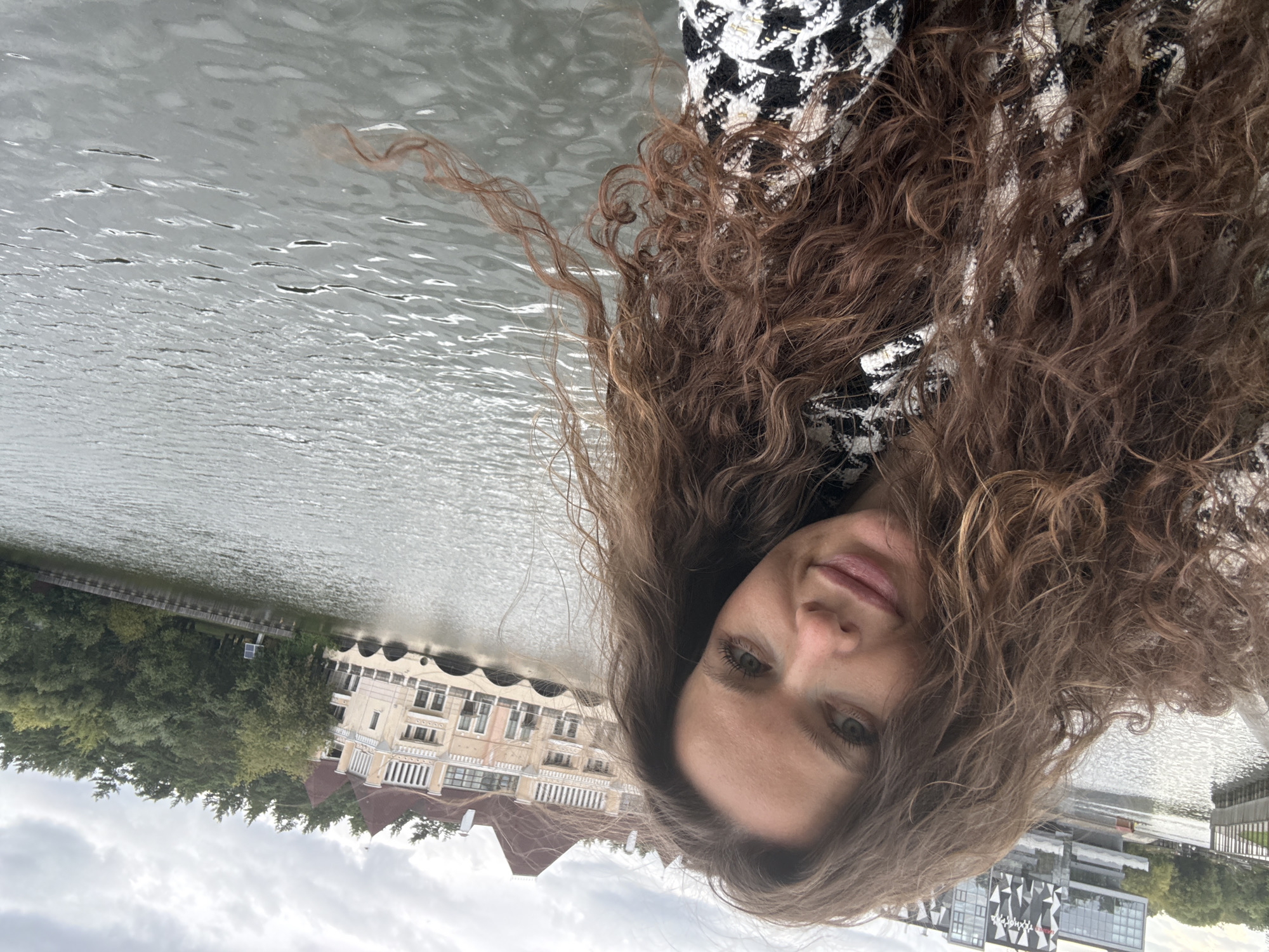 The image is a selfie of a woman with long, curly brown hair, looking towards the camera. She is wearing a black and white patterned jacket. Behind her, a body of water, likely a river or canal, reflects the cloudy sky. In the background, a large, ornate building with a red roof and arched lower levels stands on the right side of the water. To the left, a modern building with a geometric facade is visible, situated near a railing and a body of water. The setting appears to be an urban or semi-urban environment in Moscow, Russia, possibly near a park or recreational area along the waterfront. The overall atmosphere is overcast and calm.