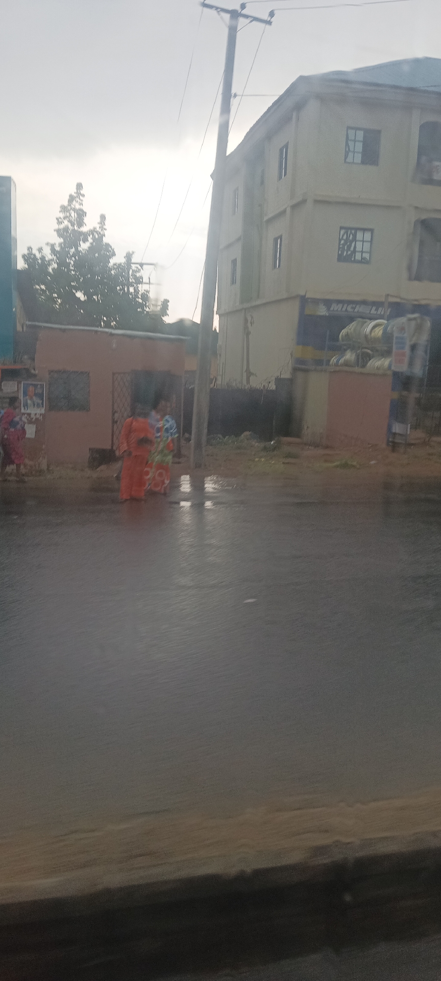 The scene is an outdoor view in Jos, Nigeria. In the foreground is a water-covered street, potentially due to rain. Three people stand near a small building with a poster. Further back, a multi-story building has a sign for "MICHELIN" along with tires displayed. A power pole is visible in the sky. The sky is overcast.