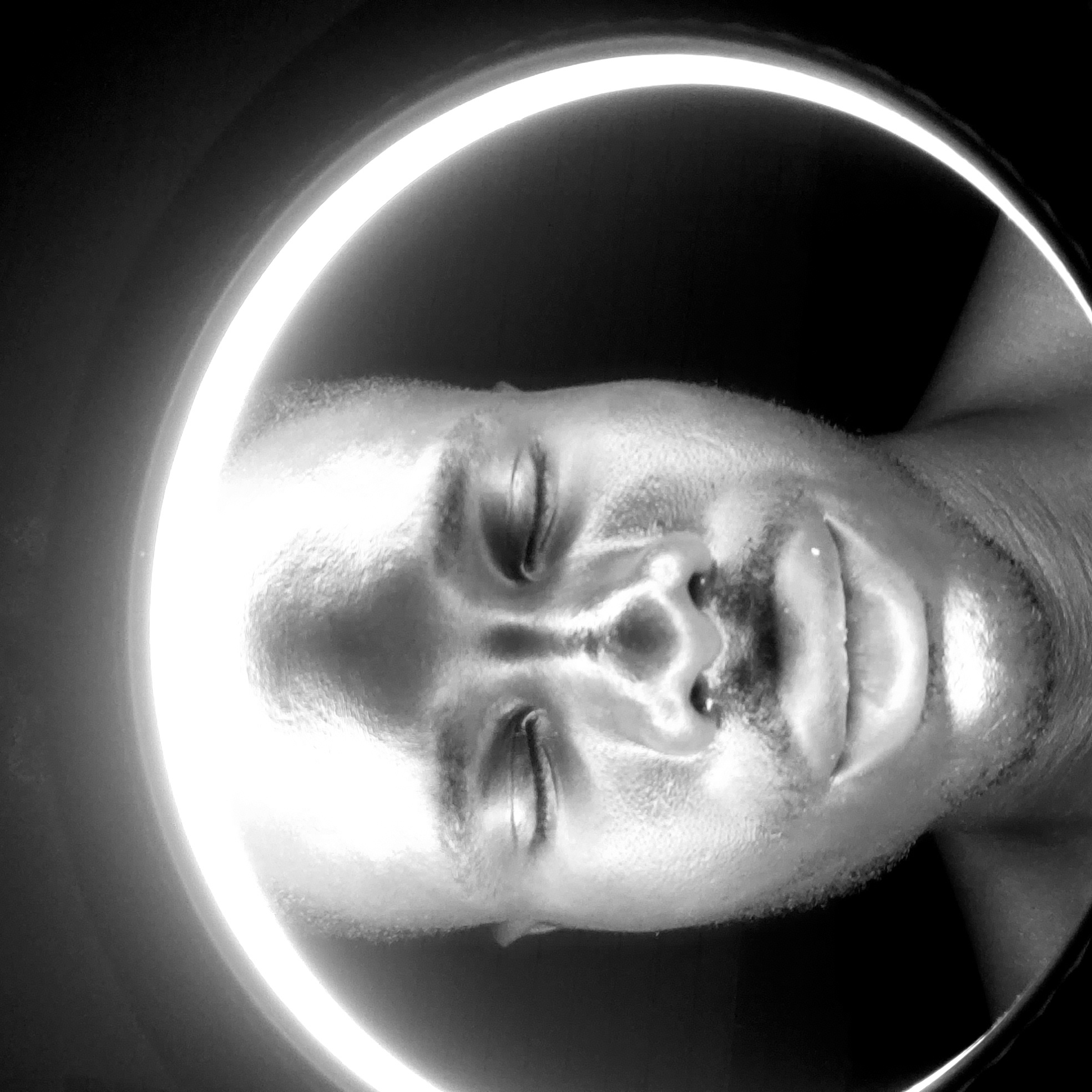 A black and white, close-up image shows the face of a man with closed eyes. The subject is partially illuminated by a bright, circular light. The environment is black and appears to be indoors. There is no indication that the image was taken in Narayi, Nigeria.