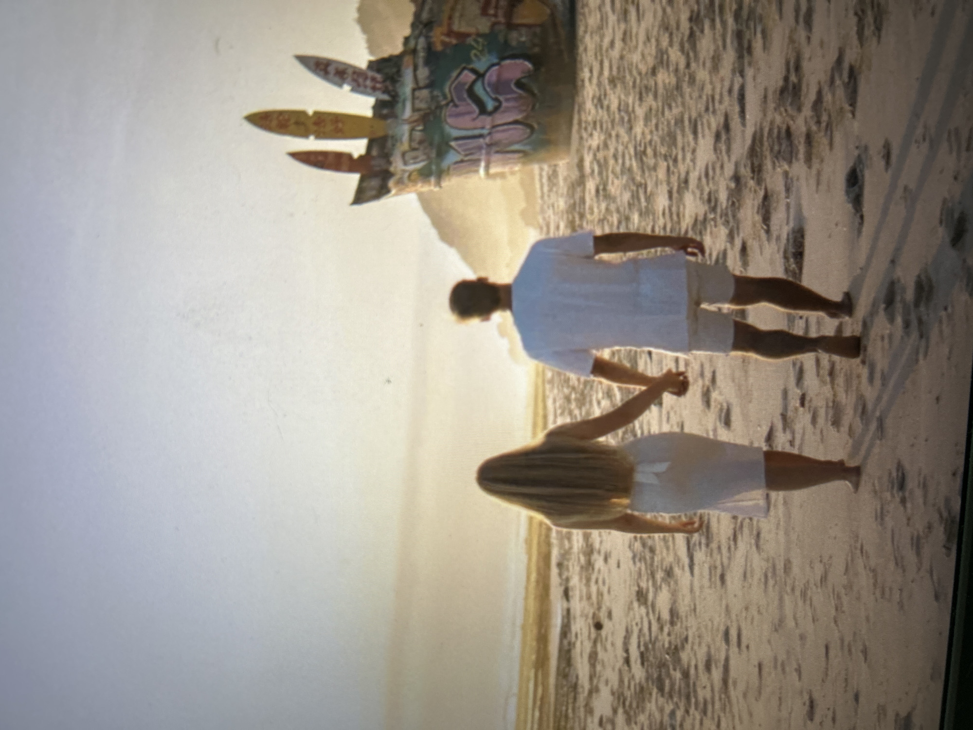 A photo shows a couple walking on a sandy beach. The sky is bright with a setting sun illuminating the scene with warm light. The beach extends to the horizon, with a blurred edge of water. In the background stands a sculpture of a tall, graffiti-covered monument. The couple, dressed in light-colored attire, hold hands as they walk together. The man wears a short-sleeved shirt and shorts, while the woman wears a short dress. The location is likely around the Uluwatu area, known for its beaches and surf culture, though the exact monument would need further investigation to pinpoint the specific beach.
