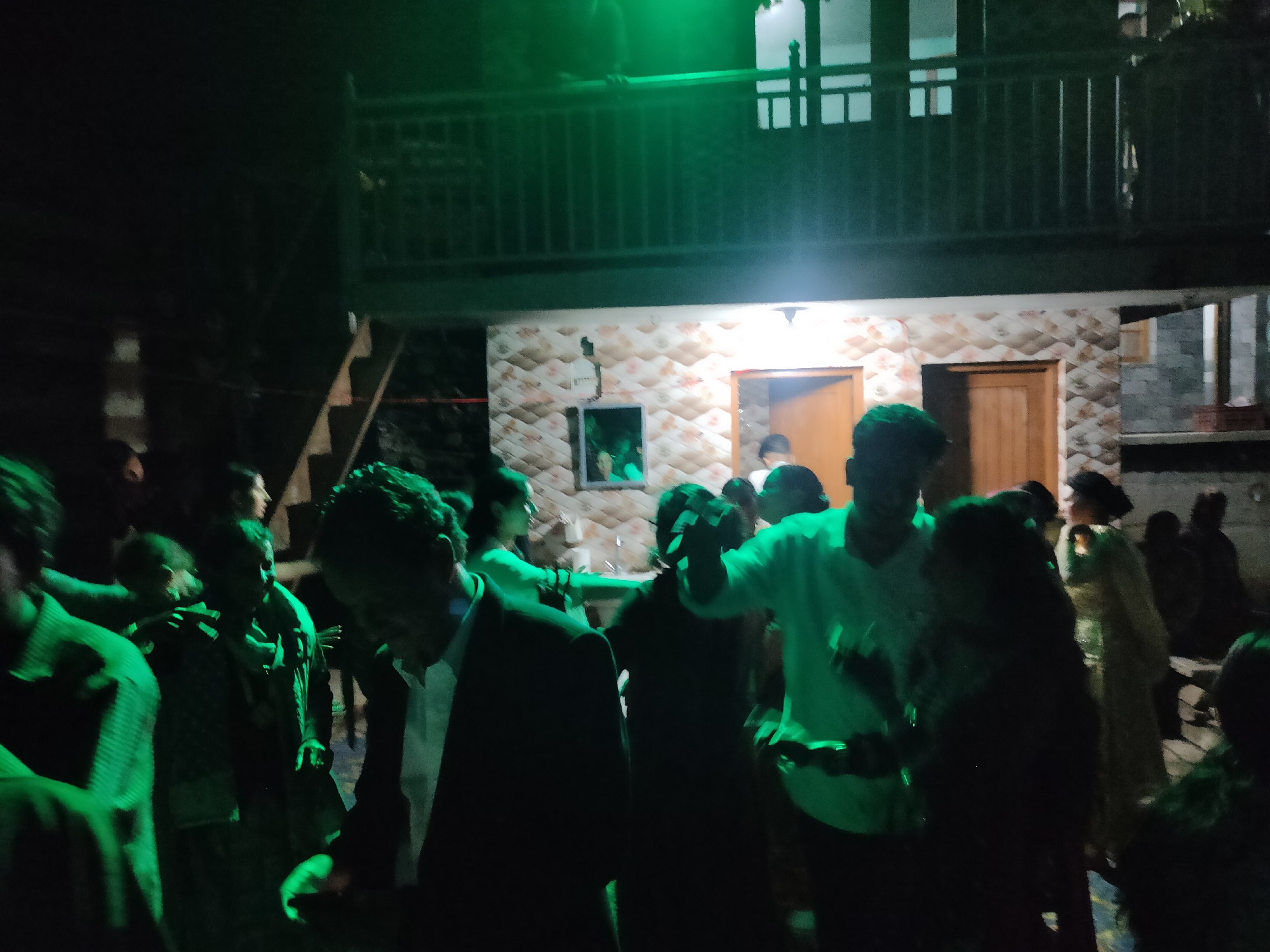 The image depicts a nighttime gathering of people outdoors, likely a party or social event. The scene is illuminated by green lighting, casting a green hue over the individuals and their surroundings. In the background, a building with a balcony is visible. The building appears to be constructed of brick and has several windows. The lower part of the building has a tiled facade. People are seen standing and interacting, some appearing to be dancing or conversing. The overall atmosphere suggests a lively and informal event. The location is identified as Damadi, India.