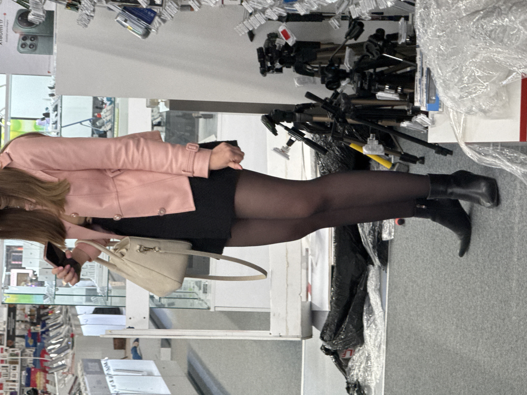 A woman stands in a store aisle. She is wearing a pink jacket, black dress, black tights, and black boots. She is holding a light beige purse and a smartphone. The store aisle contains photography equipment such as tripods. The aisle also contains electronics such as mobile phones in packaging. The exact location in Sofia, Bulgaria cannot be determined from this image.