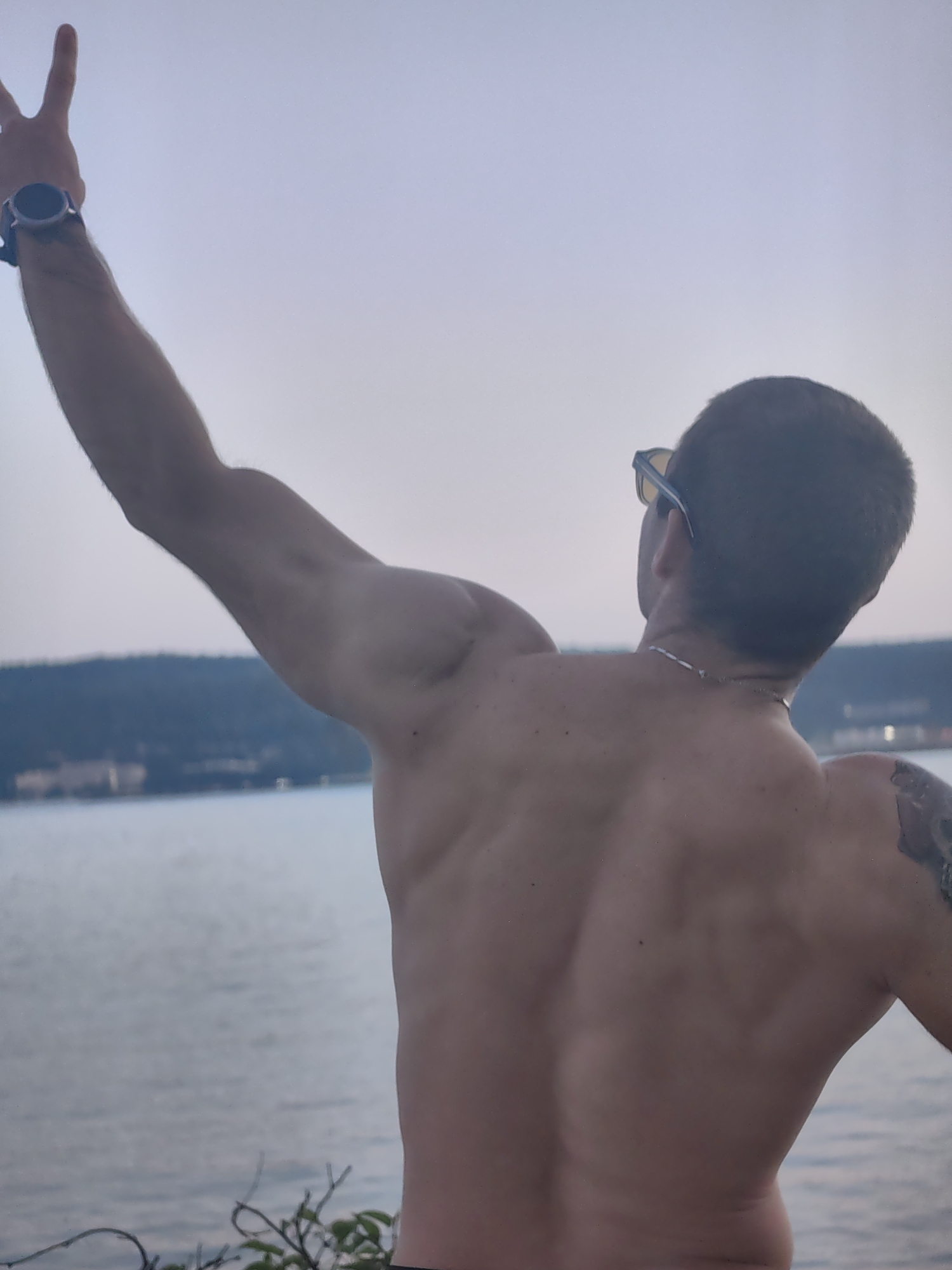 A shirtless man stands with his back to the camera, facing a body of water. His left arm is raised, with his index finger and middle finger extended in a peace sign. He is wearing a watch on his left wrist. He has sunglasses on and a chain around his neck. In the background, a blurred treeline runs along the shore of the water, suggesting a natural or park-like setting. The sky is overcast. Based on the context, this image could be taken at the Varna Sea Garden, a large park in Varna, Bulgaria, which features extensive coastlines and natural scenery.