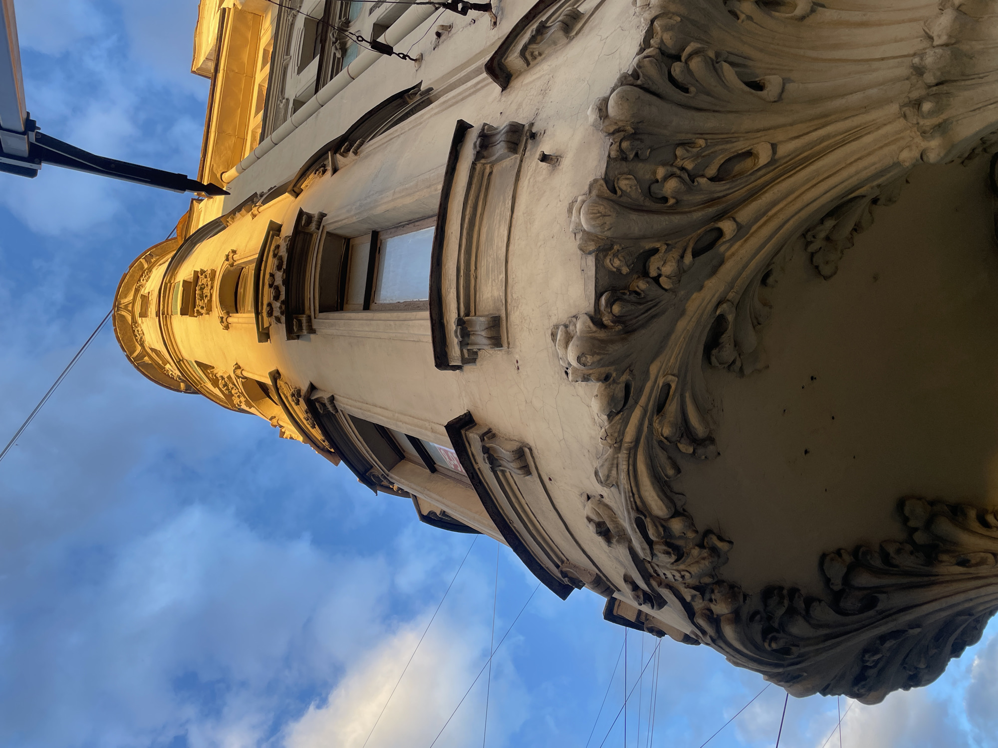 This is a low-angle, eye-level, close-up shot of the ornate corner of a historic building in Saint Petersburg, Russia, bathed in the warm, golden light of the setting sun. The building's facade is adorned with intricate sculptural details, featuring floral motifs and elaborate moldings on a curved bay window. Above, a bright blue sky is visible, scattered with soft white clouds. Overhead, electrical wires crisscross the frame, indicating an urban setting. The architectural style suggests it could be located in the historic city center, perhaps in an area known for its Art Nouveau or pre-revolutionary architecture. No people are visible in the frame.