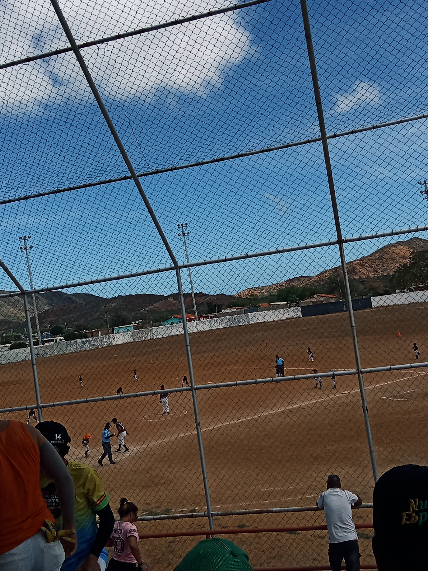 The image shows a baseball game in progress, viewed from behind a chain-link fence, likely from a spectator stand. The baseball field is brown dirt, and the back wall of the field is visible, bordered by white concrete and additional fencing. The sky is bright blue with some puffy white clouds. In the background are dry, tan hills.
Several people are visible on the field, including players and an umpire. At least two players are engaged in what appears to be a play at home plate. Spectators are visible along the bottom edge of the image, including individuals in an orange shirt, yellow shirt with a number 14 on a hat, pink shirt and one wearing a hat with "Nue Esp" embroidered on it.
The scene is lit by bright daylight. The location is assumed to be Juan Griego, Venezuela, based on the prompt.