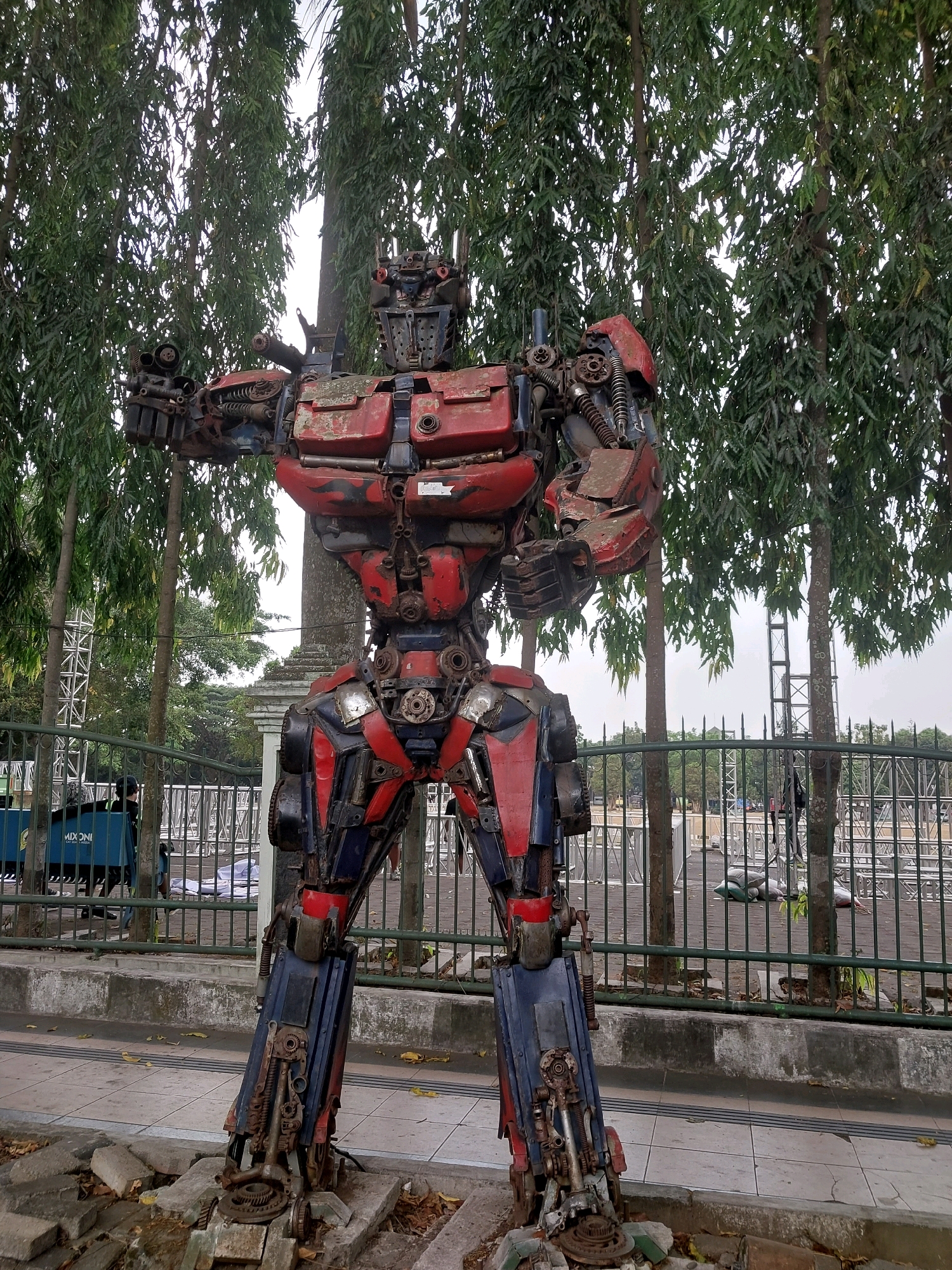 A large, weathered, red and blue robot sculpture, resembling Optimus Prime, stands in front of a row of trees and a metal fence. The robot is constructed from scrap metal and automotive parts. Its right arm is raised, and its left arm is bent at the elbow. The robot is positioned on a sidewalk with broken concrete pieces at its base. In the background, beyond the fence, are scaffolding structures and a few people. The scene appears to be in an outdoor public space in Malang, Indonesia. The sky is overcast.