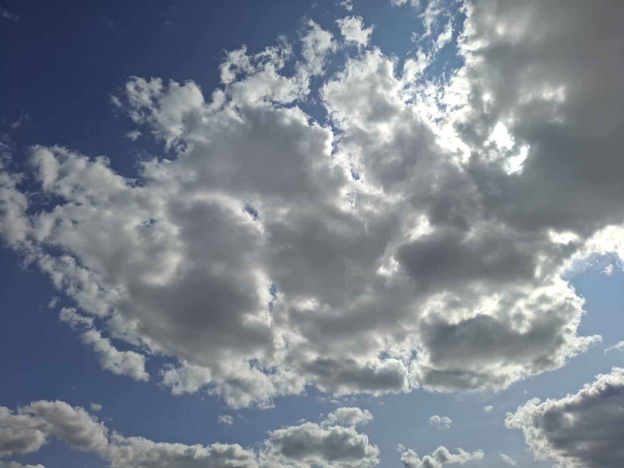 The image depicts a sky filled with a mix of white and dark gray clouds against a bright blue sky. The sunlight filters through some of the clouds, creating bright patches and highlights. The clouds are varied in size and texture, with some appearing fluffy and others more dense and ominous. The overall impression is one of a dynamic sky, possibly indicating changing weather conditions. The image does not contain any specific landmarks or details that would allow for pinpointing a location within Chorna, Ukraine.