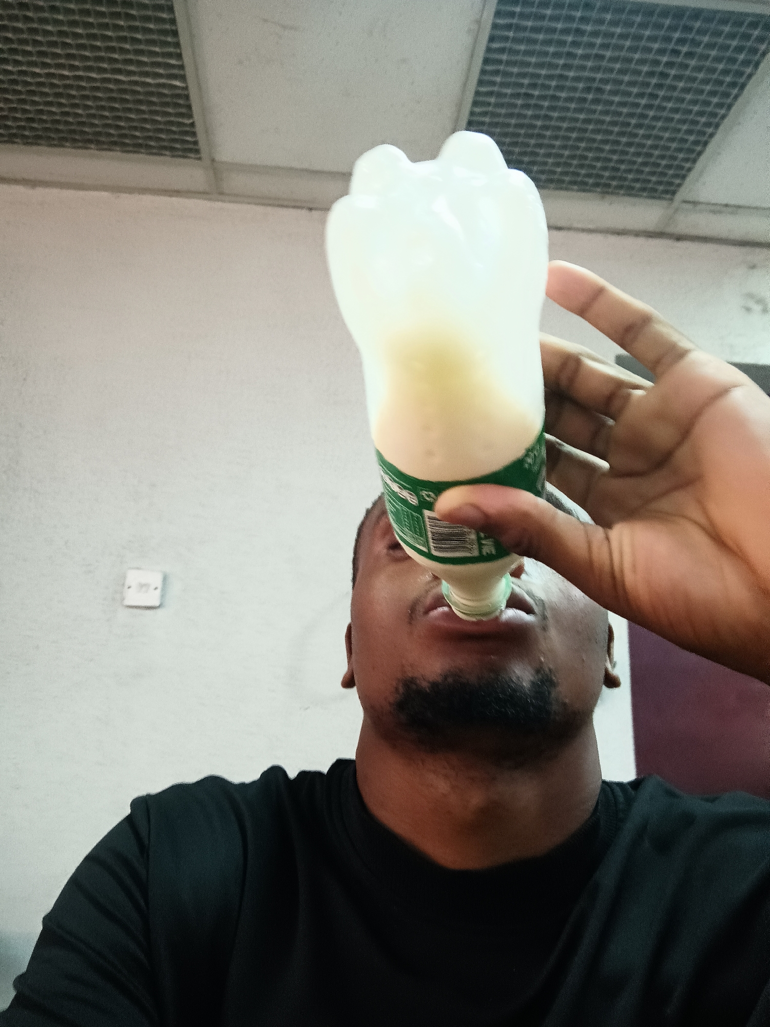 A close-up, low-angle indoor shot captures a young man, likely of African descent, drinking from a clear plastic bottle. He wears a black crew-neck shirt and has a short goatee beard. His head is tilted back as he brings the bottle to his mouth, obscuring most of his face. His right hand firmly holds the bottle, which contains a milky white or light yellow liquid. The bottle has a distinct four-lobed top and a green label displaying a barcode and small, illegible white text.
The setting appears to be an indoor space in Kakuri Gwari, Nigeria. The background features a light-colored wall, possibly off-white or light grey, with a white electrical switch or outlet visible on the left. Above the wall, a ceiling with a dark rectangular grid pattern, suggesting a ventilation system or acoustic panels, extends across the upper part of the frame. A darker, possibly purple or brown, surface is partially visible on the right side of the image. The lighting indicates an indoor daytime scene.