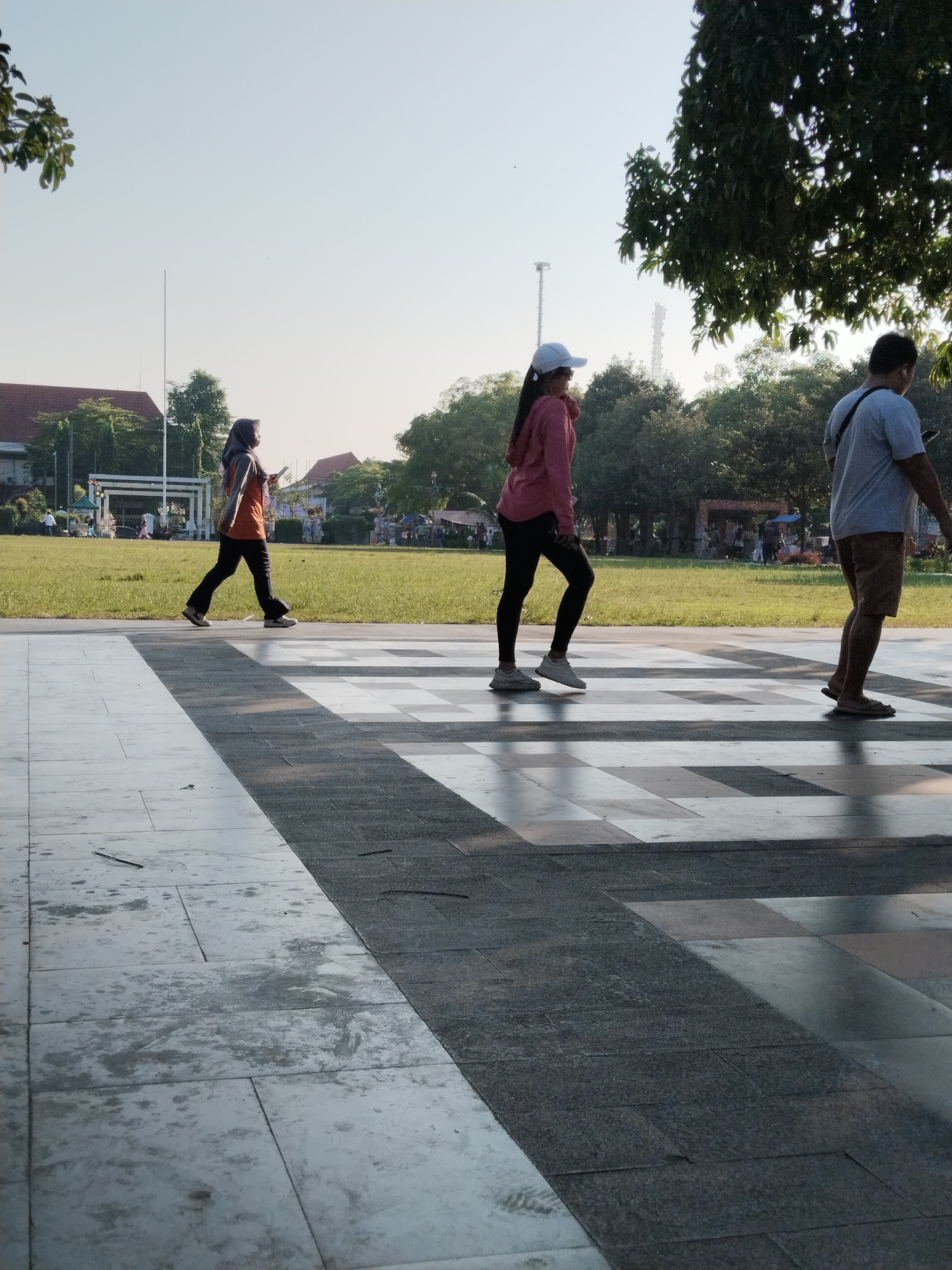 Three people are walking across a paved area in a park-like setting. The person on the left is wearing a headscarf and an orange top with black pants, and is looking at their phone. In the middle is a person wearing a pink hoodie, black leggings, and a white cap, looking to their right. On the right is a person wearing a light blue t-shirt, brown shorts, and flip-flops, looking to their left. Behind them, a grassy field with trees and buildings can be seen. The sky is clear and bright, suggesting it is daytime, likely morning or late afternoon due to the long shadows. The overall atmosphere appears relaxed and leisurely, with people enjoying the outdoor space. There is no visible text in the image.