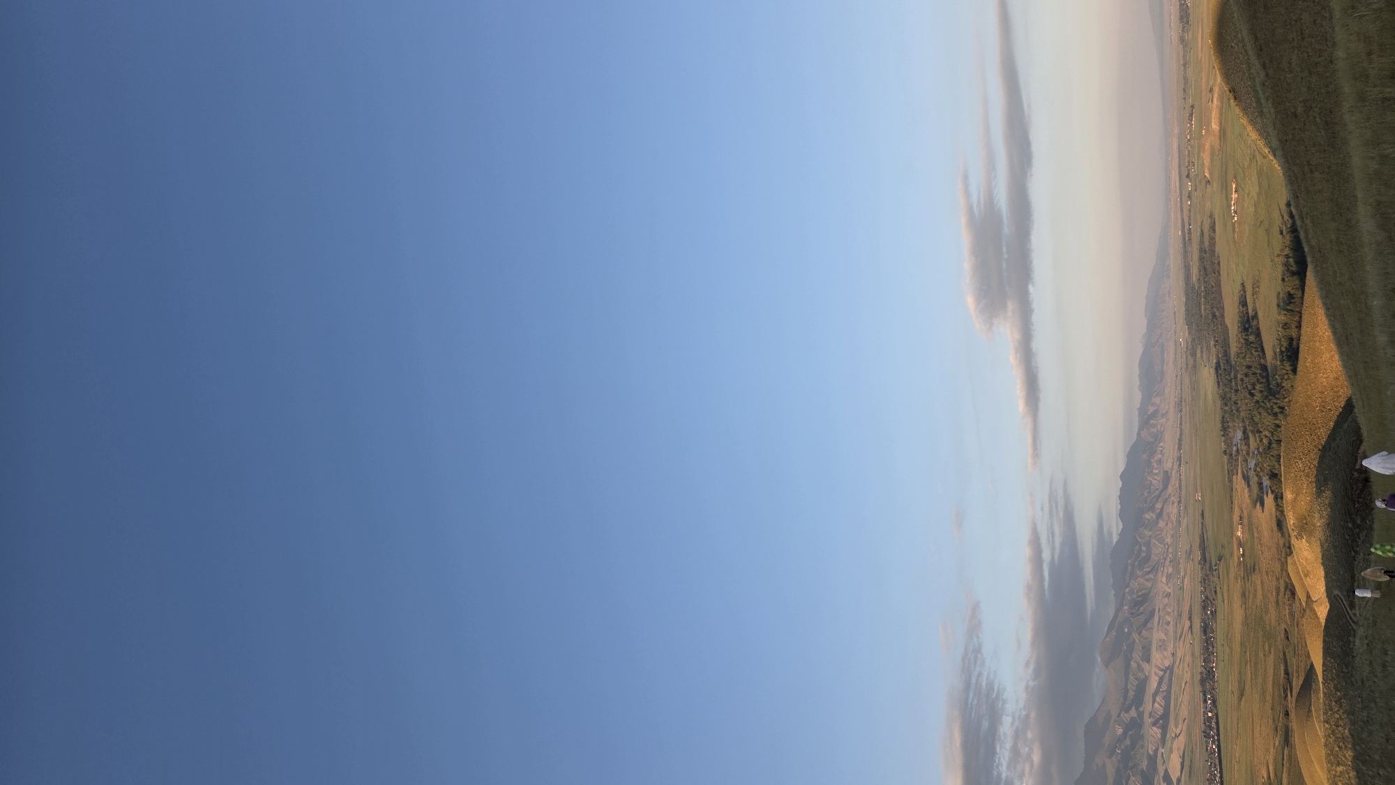 The image depicts a panoramic view of a valley near the city of At-Bashi, Kyrgyzstan, under a clear blue sky. In the foreground, rolling hills covered in dry grass are illuminated by the setting sun, casting long shadows. A group of five individuals, dressed in white or light-colored garments, are walking down one of the hills. In the mid-ground, the valley floor is a patchwork of green fields, a winding river, and scattered trees. A small town, likely At-Bashi, is visible on the left side of the valley, with buildings clustered together. In the background, a range of majestic, rugged mountains stretches across the horizon, their peaks softened by the haze of distance and the golden light of dusk. Wispy clouds are scattered across the sky, particularly in the upper left and right portions of the image. The overall scene conveys a sense of peaceful rural landscape bathed in the warm glow of the evening.