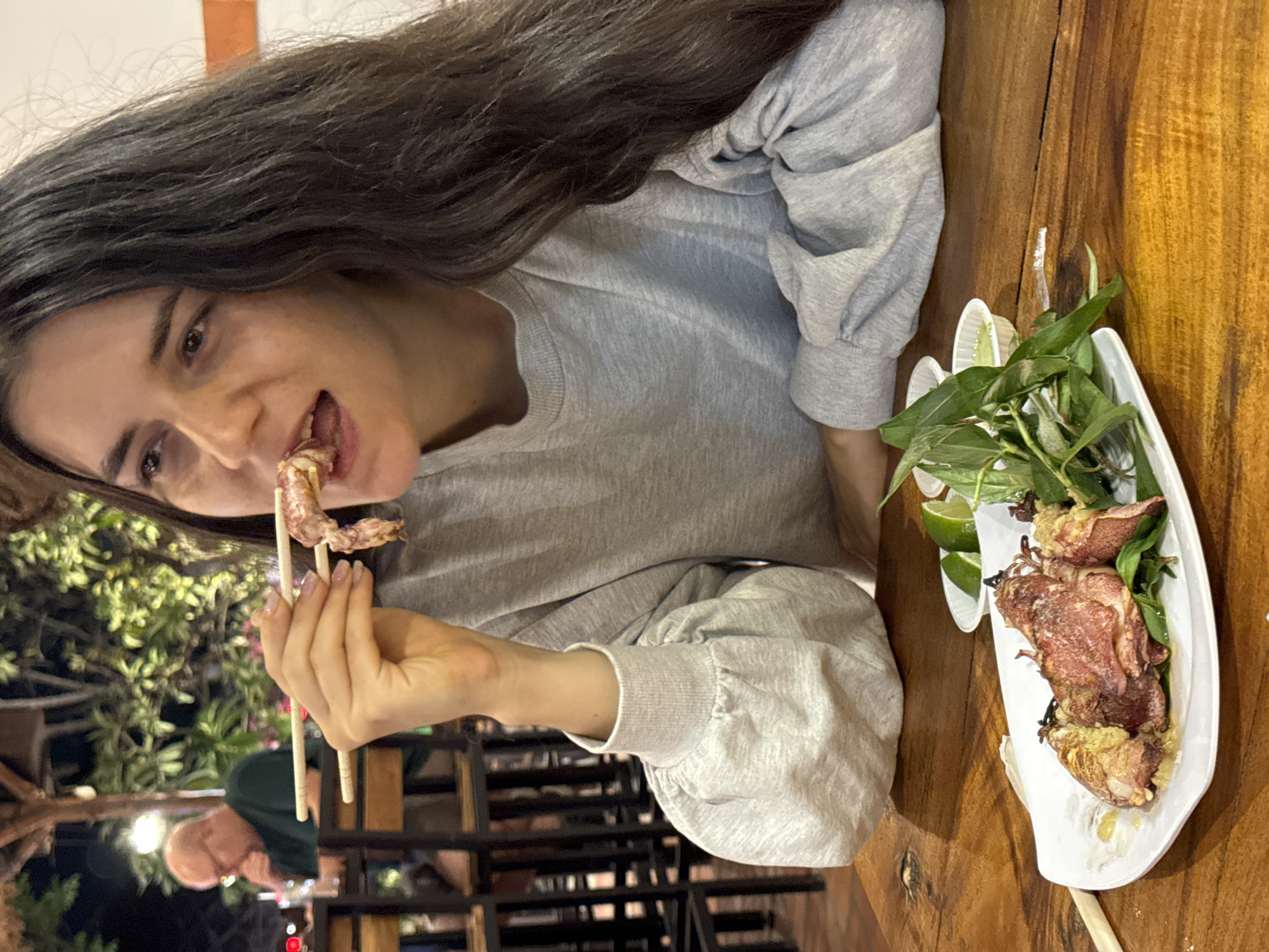 A young woman with long, dark hair, wearing a light grey sweatshirt, is captured mid-meal in an image that is rotated 90 degrees counter-clockwise. She is looking towards the viewer with a slight smile, her mouth open as she brings a piece of food, likely grilled squid, to her mouth using wooden chopsticks. The food held by the chopsticks appears to be part of a larger dish served on a white rectangular plate situated to her right on a rustic wooden table.
The plate features several pieces of whole grilled squid, which appear golden-brown and slightly charred, indicating they have been recently cooked. The dish is garnished with fresh green herbs, possibly basil or mint, and bright green lime wedges, suggesting a Vietnamese culinary setting, consistent with the Mũi Né location context.
The scene appears to be an outdoor or semi-outdoor restaurant at night. The background is blurred but shows dark foliage and soft, warm artificial lights, creating a relaxed evening ambiance. Another individual, a man, is dimly visible and out of focus in the deeper background. The woman's expression conveys contentment and enjoyment of her meal. There is no visible text in the image.