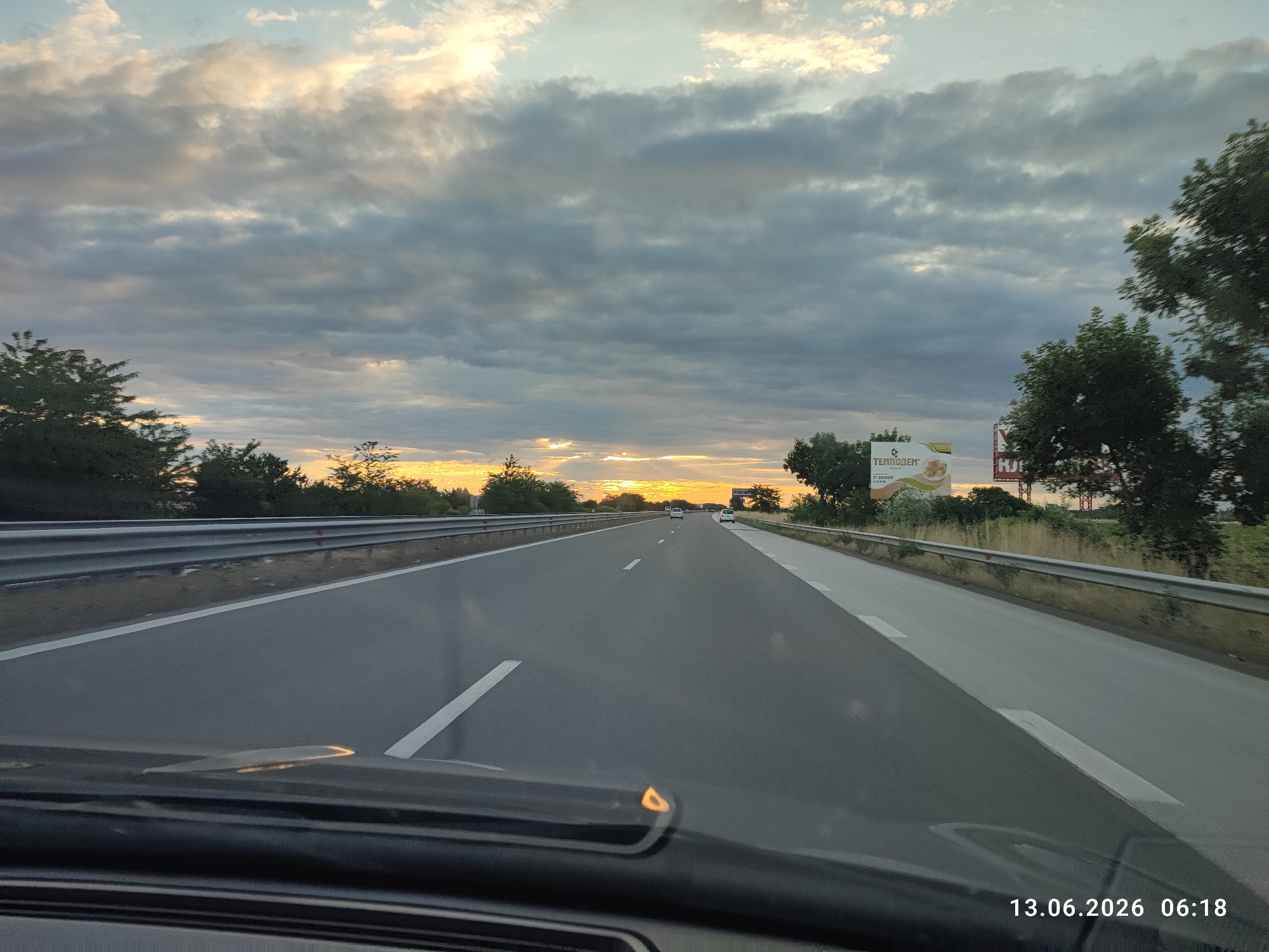 The image captures a view from a vehicle driving on a highway towards a sunset. The sky is filled with dramatic, dark clouds interspersed with patches of bright light from the setting sun, creating a dramatic and somewhat ominous atmosphere. The highway itself is paved, with clear white lane markings. Guardrails line the sides of the road. On the right side of the highway, there are trees and vegetation, and a billboard is visible. The timestamp "13.06.2026 06:18" is visible in the lower right corner of the image. The location of Tsaratsovo, Bulgaria is not explicitly visible in the image, but it is suggested by the context provided with the prompt.