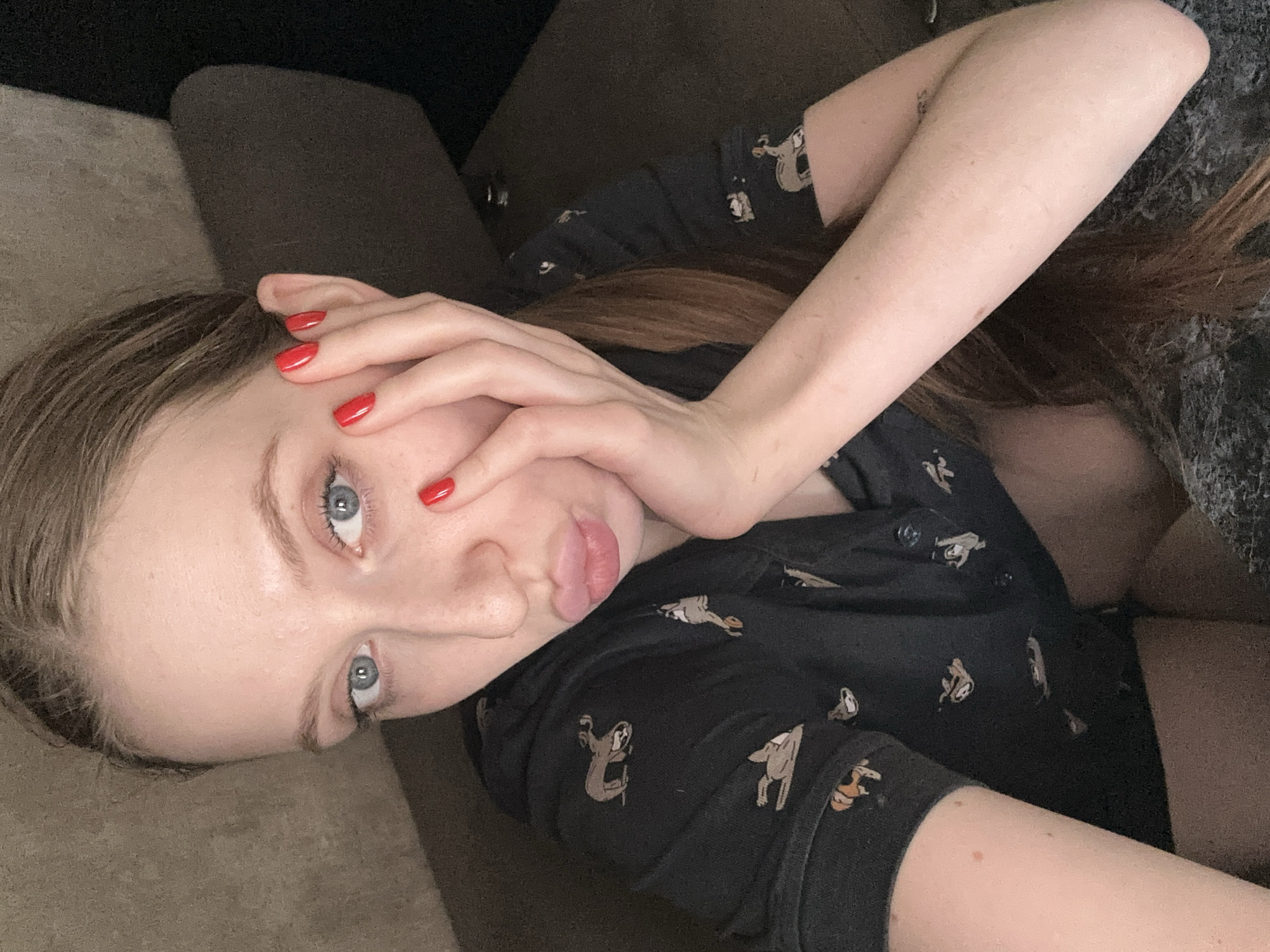 A young woman is depicted in a close-up, slightly tilted portrait, lying down indoors. She has light skin, striking blue eyes, and light brown hair. Her nails are painted a vibrant red. One hand is positioned near her face, with fingers lightly touching her forehead and cheek. Her lips are slightly parted in a subtle pout.
She is wearing a dark, possibly black, short-sleeved button-up shirt featuring a pattern of small, light-colored animals, which appear to be sloths or similar creatures. The background is dark and indistinct, suggesting she is resting on a sofa or bed. The lighting in the scene is soft and dim, indicating an indoor setting, likely in the evening or at night. The woman is looking directly towards the viewer with an expressive gaze. There are no specific location cues visible in the image itself to suggest Chaikovsky, Russia, nor is any text visible. The image conveys a casual and intimate atmosphere.